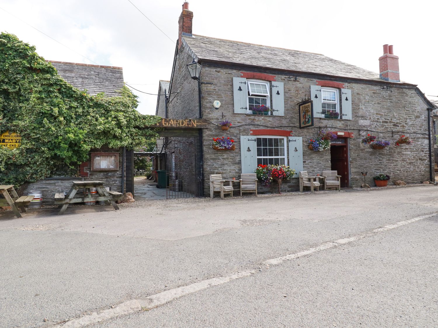 A stone building with wooden chairs and flower pots outside next to an entrance with a garden sign at Gwynn Cottage in St Mabyn