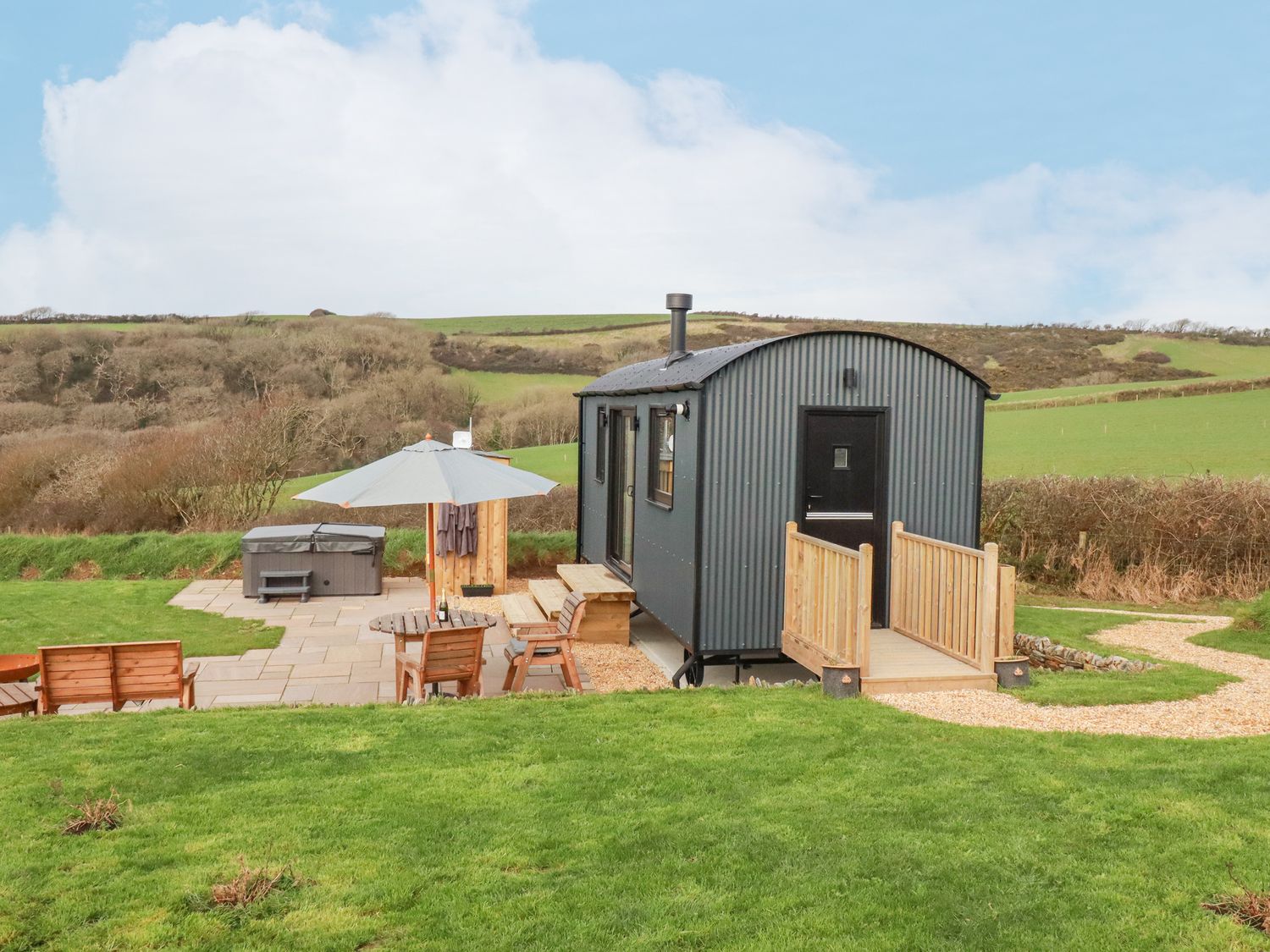 A small black corrugated metal hut with a wooden ramp and an outdoor patio area with chairs a table an umbrella and a hot tub in a grassy area at Northcott Hut in Stibb near Bude