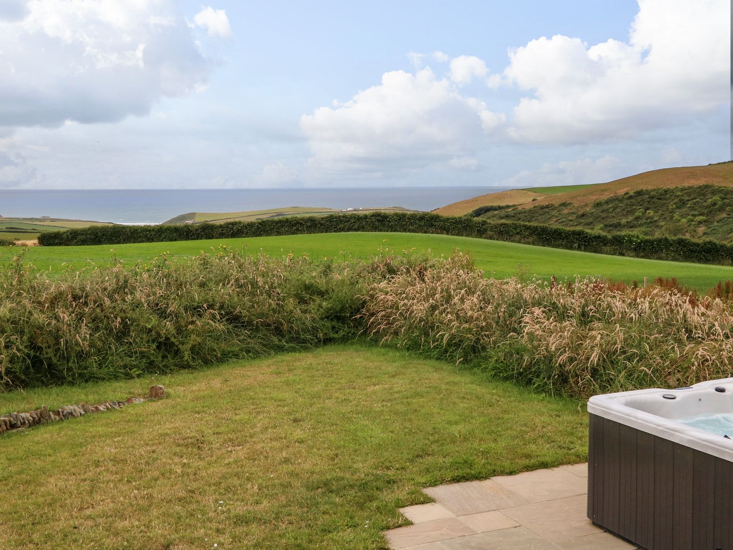 A backyard with grassy lawn and bushes with ocean and hills in the background and a hot tub in the corner at Northcott Hut in Stibb near Bude