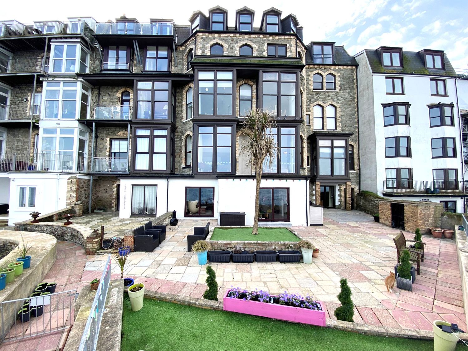 Exterior view of a multi-story building with large windows and balconies a patio area with seating planters and a tree at 1 Cheyne Beach Apartments in Ilfracombe