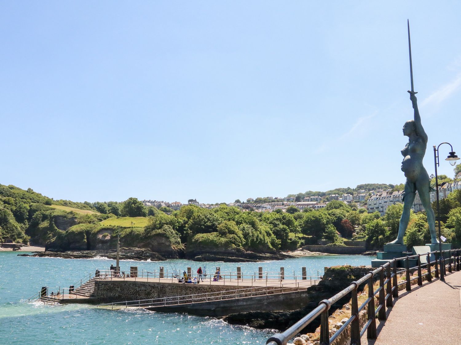 A waterfront promenade with a statue of a pregnant woman holding a sword and a pier extending into the sea at 1 Cheyne Beach Apartments in Ilfracombe