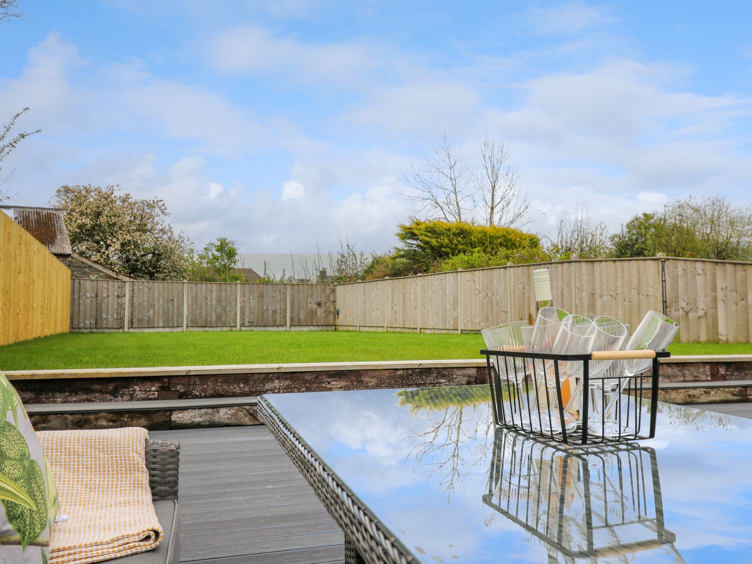 A backyard with green grass and wooden fencing with a glass table holding a basket of glasses and a bottle at 2 Penporth in Rhayader