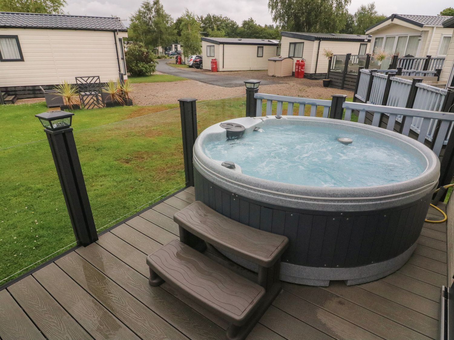 A hot tub on a wooden deck with steps surrounded by glass and railing at Waterside Lodge South Lakeland Leisure Village near Carnforth