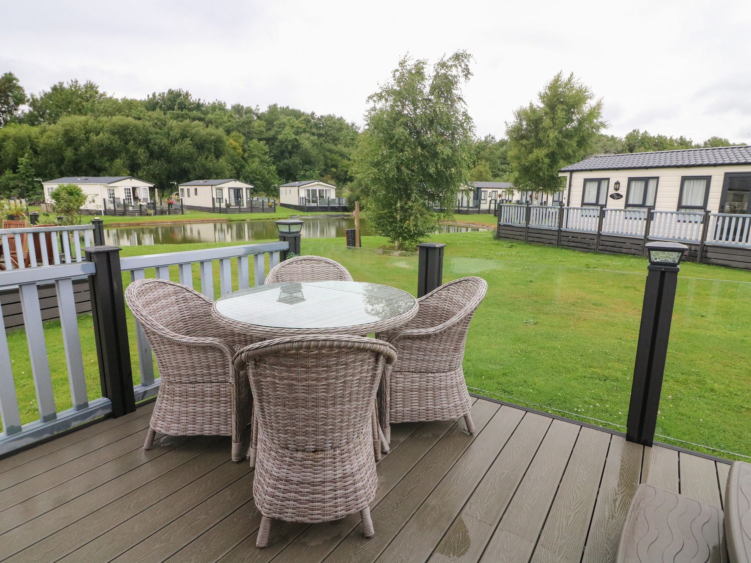 An outdoor patio with wicker chairs and a glass table overlooking a pond and several white lodges at Waterside Lodge South Lakeland Leisure Village near Carnforth