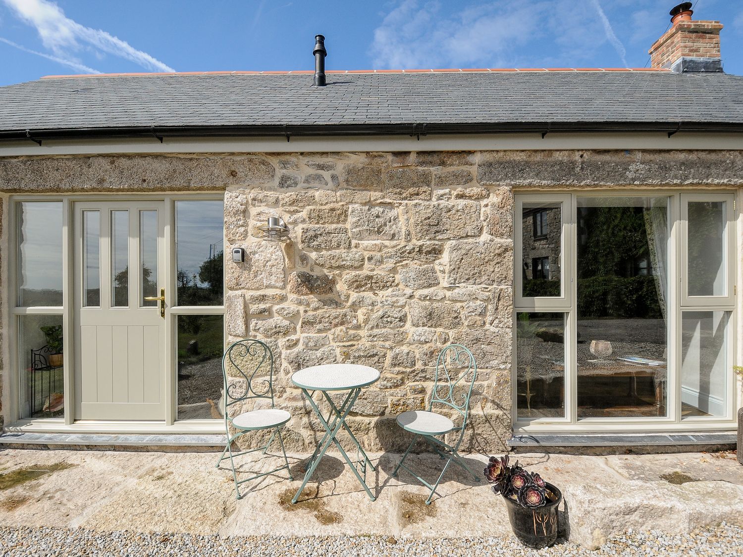 A stone house exterior with a door and large windows a small round table with two chairs and a black bucket with plants at The Forge in Mawnan Smith