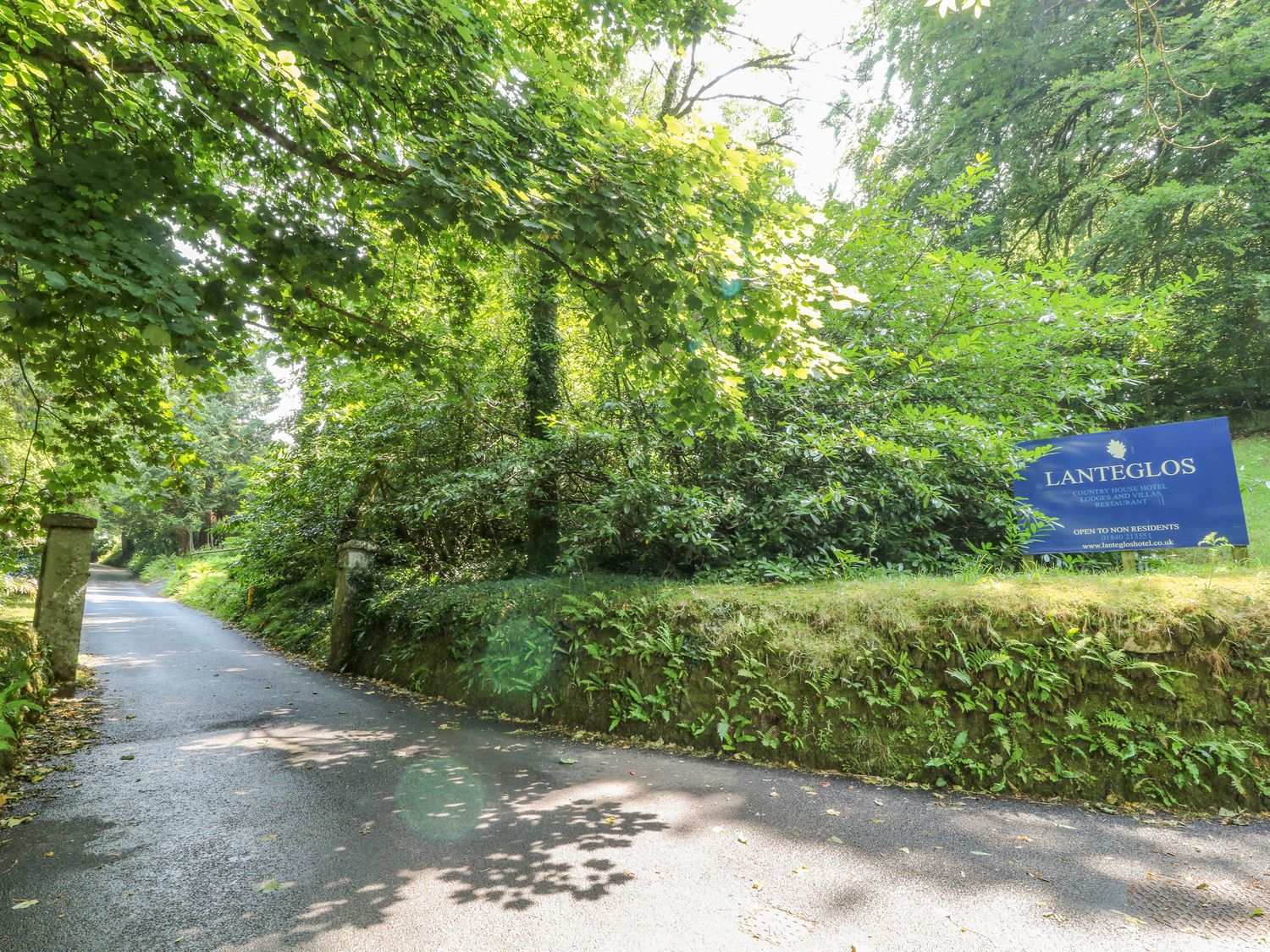 A paved road through green trees and bushes with a blue Lanteglos hotel sign on the right at Lodge 16 in Lanteglos near Camelford