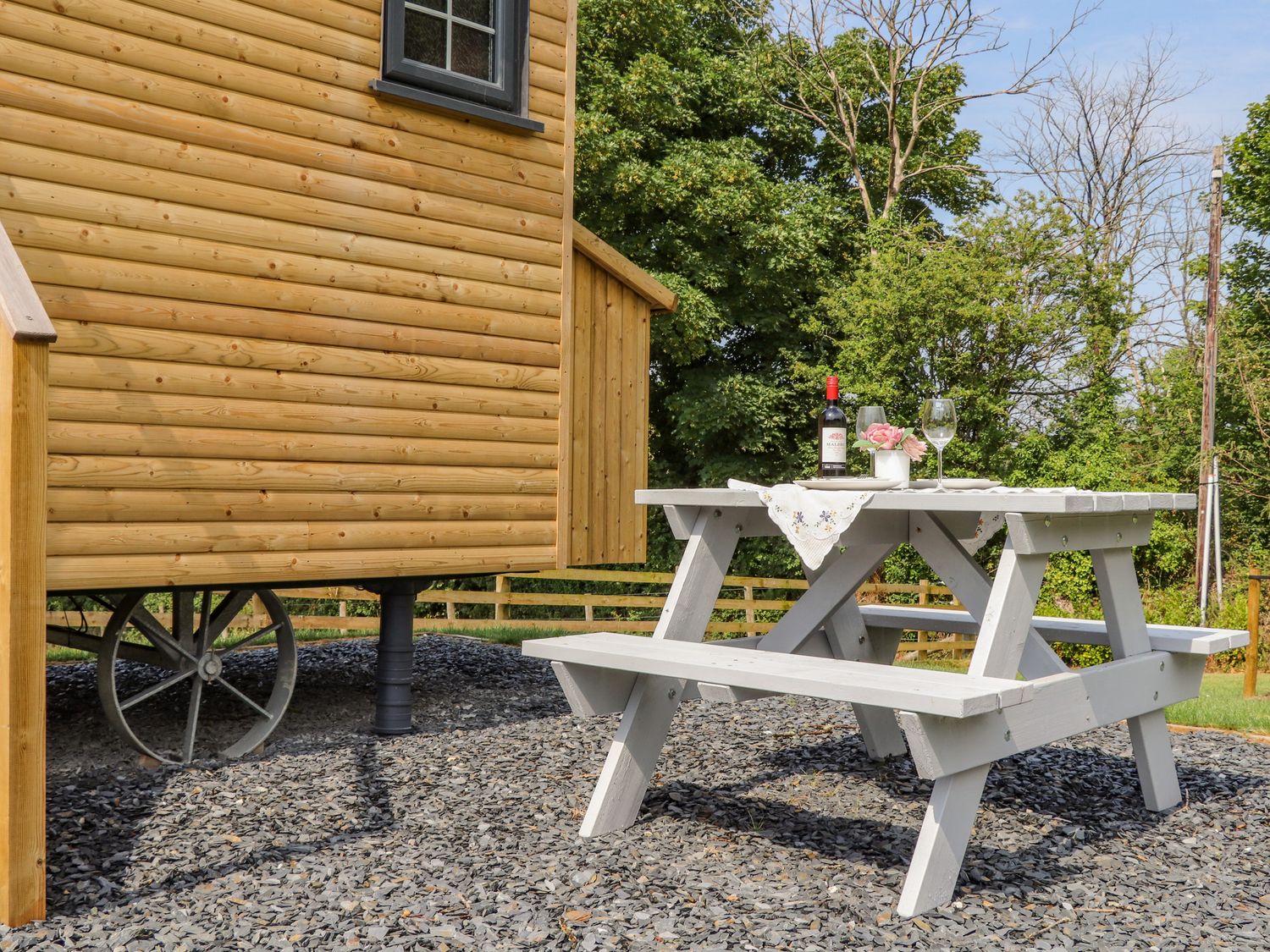 A wooden picnic table with a bottle of wine glasses and flowers next to a wooden wagon in a garden at Rose in Illogan