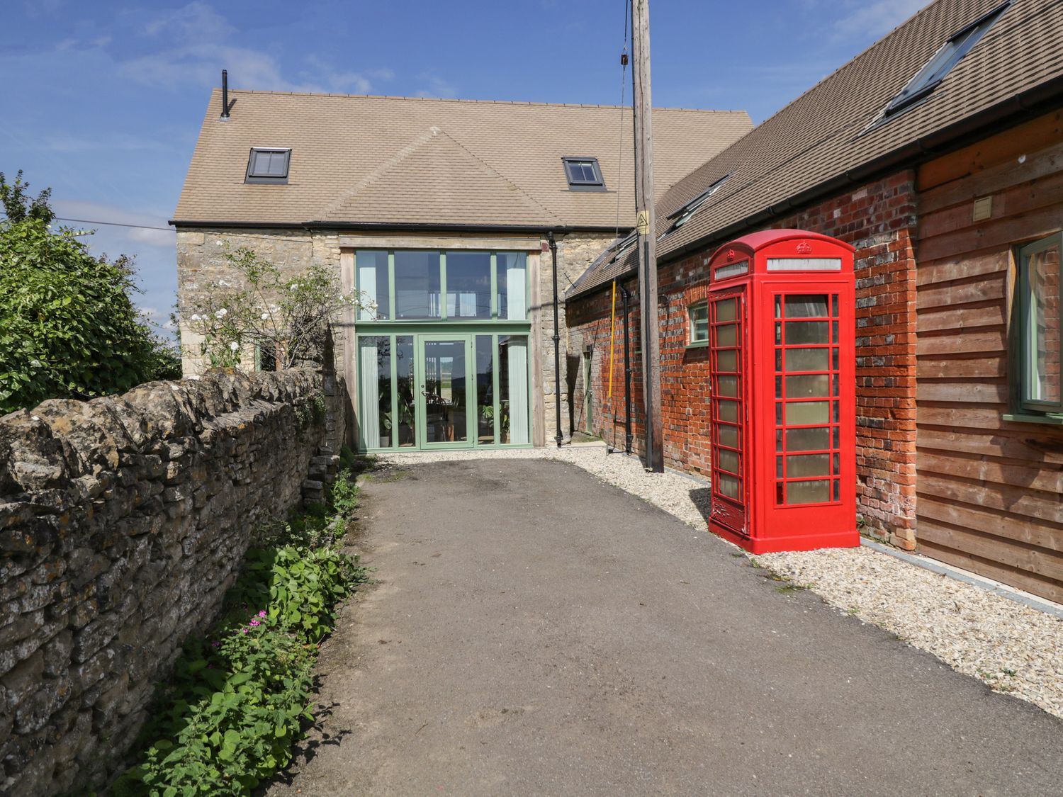 A driveway between a stone wall and a building with a red telephone box and large windows at The Old Barn in Barnard Gate near Eynsham