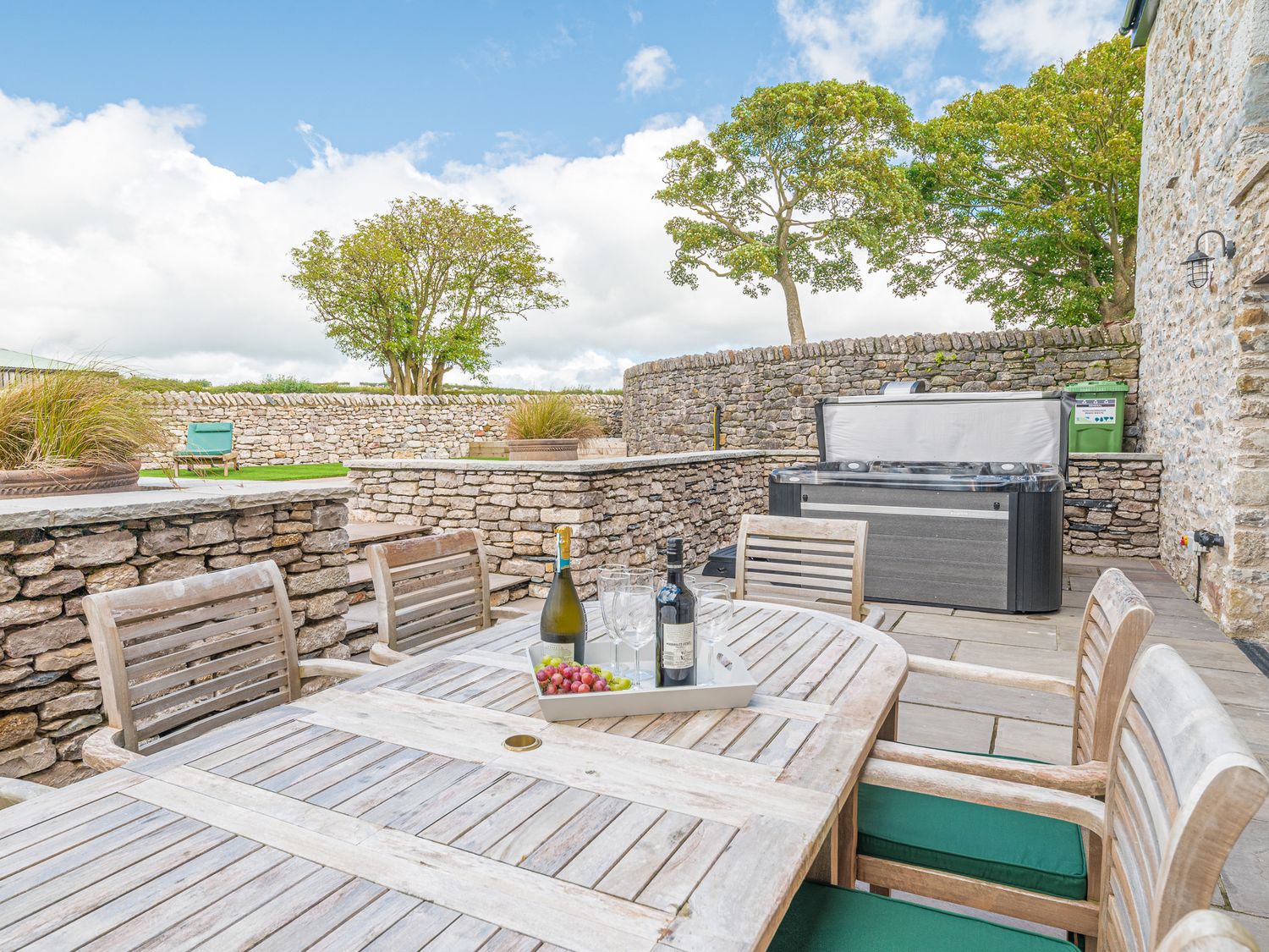 An outdoor patio with a wooden table and chairs a wine bottle and glasses on the table a hot tub and stone walls at Old Stable in Roose
