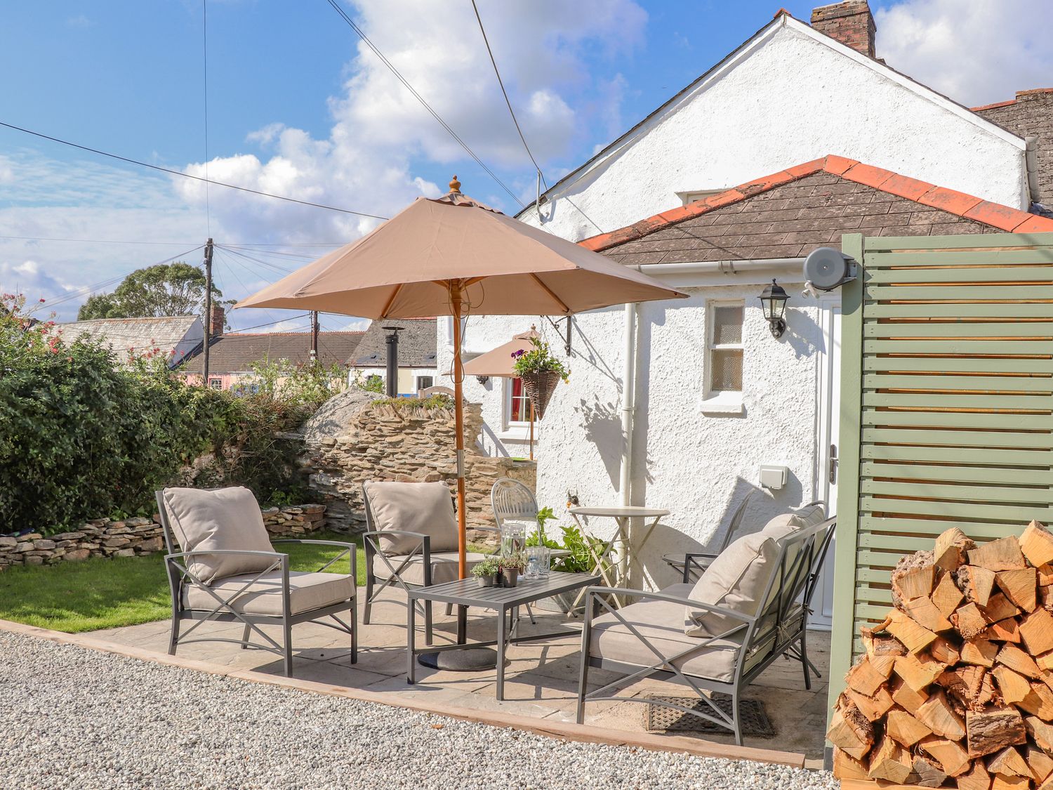 An outdoor patio seating area with chairs, a table, umbrella, and stacked firewood at Trewenna in Roseland Tregony