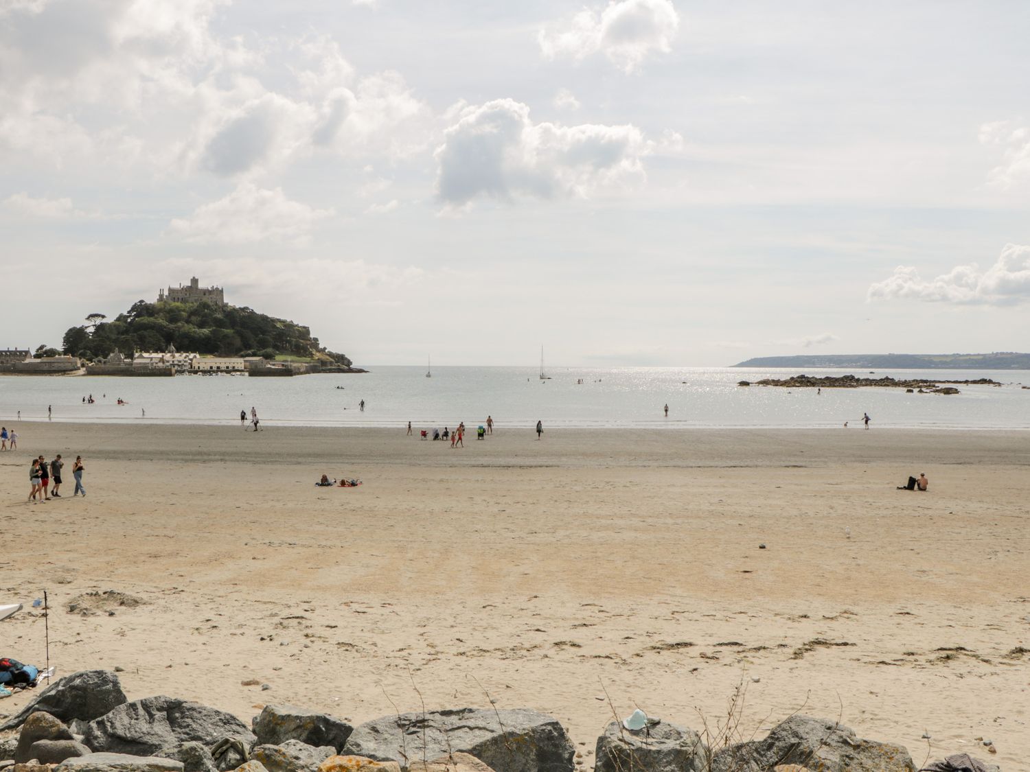 A sandy beach with people walking and sitting near the water and an island with a castle in the sea at Meledor in Marazion