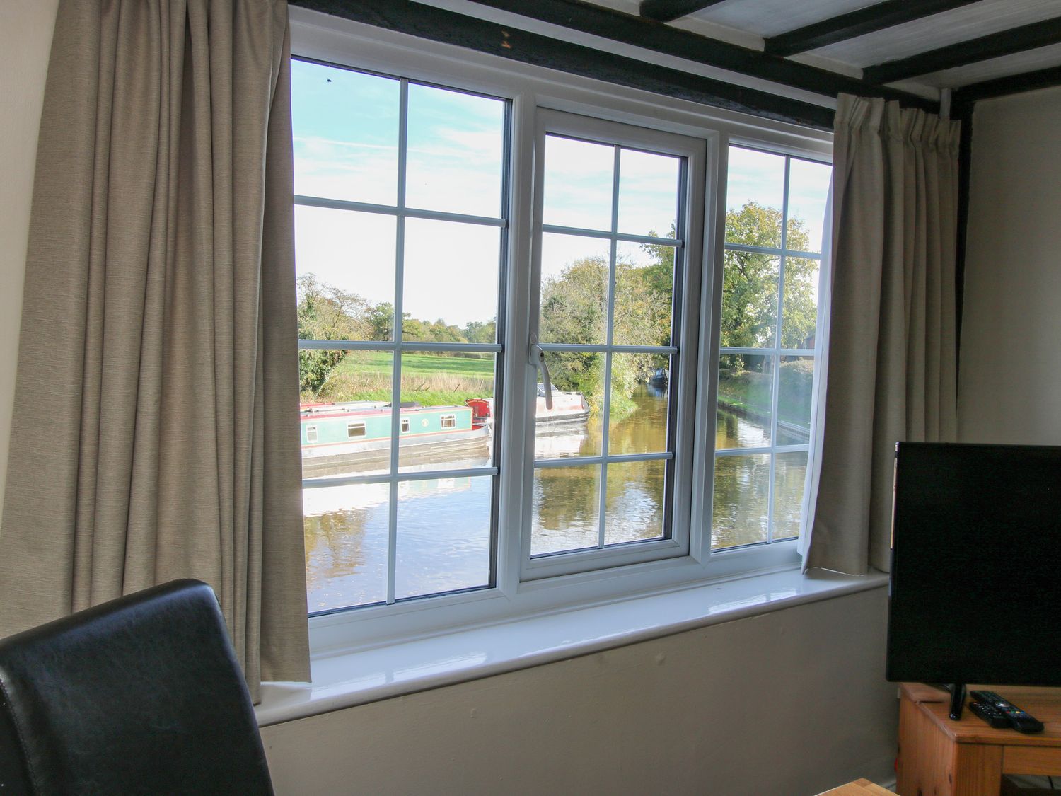 A window with beige curtains overlooking a canal with narrowboats visible from inside a room at Mill End in Wrenbury