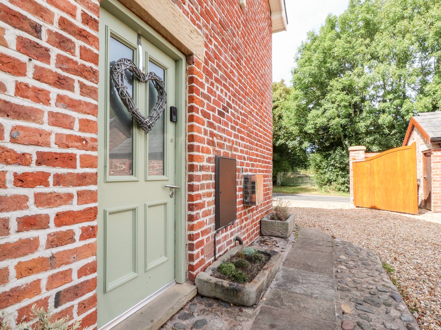A green door with a heart-shaped wreath on a red brick building next to a pathway and a wooden gate at Noah's Barn in Hambleton Lancashire