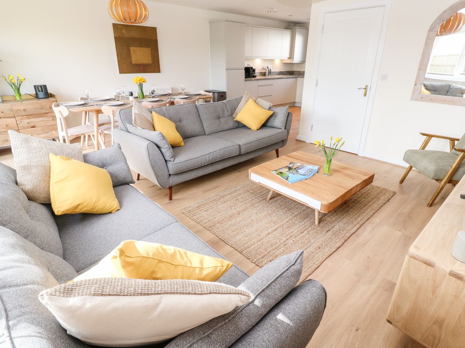A living room with two grey sofas with yellow and beige cushions a wooden coffee table with magazines and flowers a dining table set with plates and glasses and a wooden sideboard at The Bright House in St Eval near St Columb Major