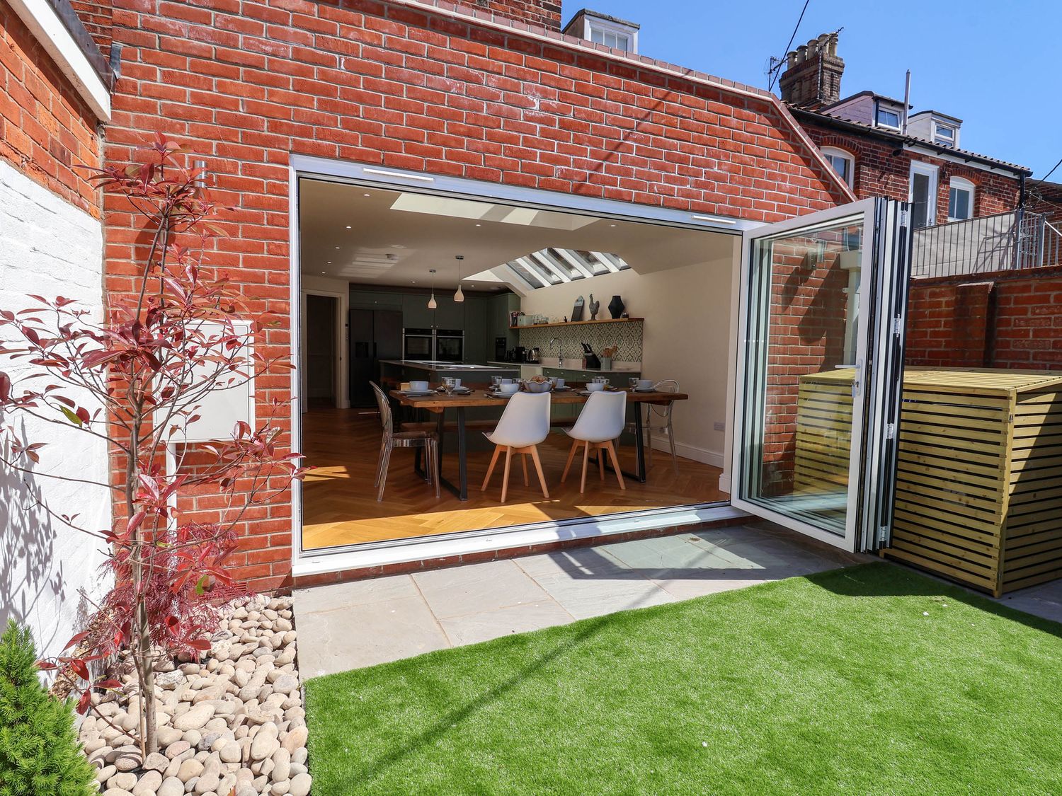 A dining area with a wooden table and chairs inside a house with large folding glass doors opening to a small garden with artificial grass at Lighthouse View Southwold