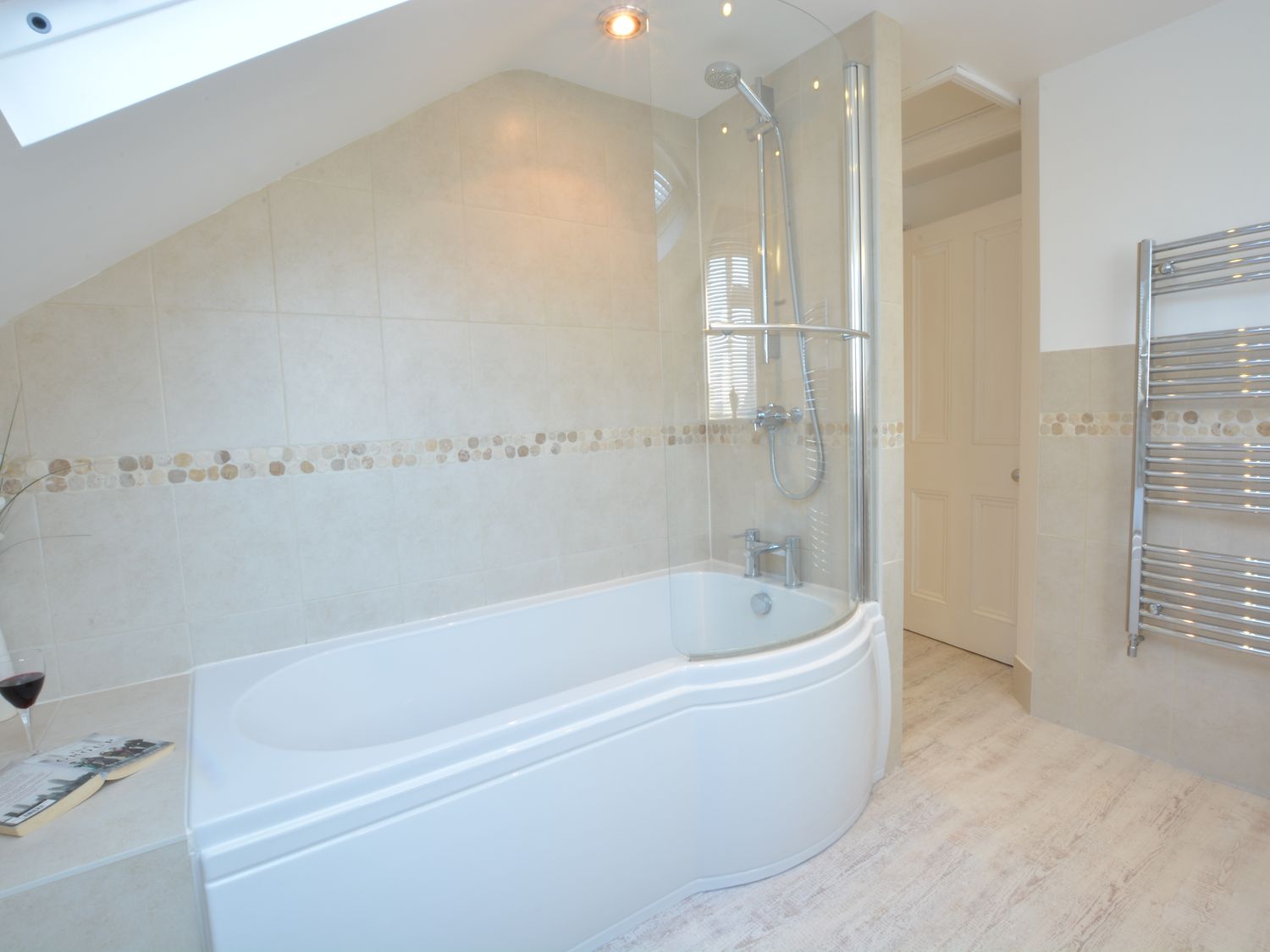 A bathroom with a bathtub and shower enclosure a glass screen a towel radiator and a book and glass of wine on the ledge at The Southwold Loft in Southwold