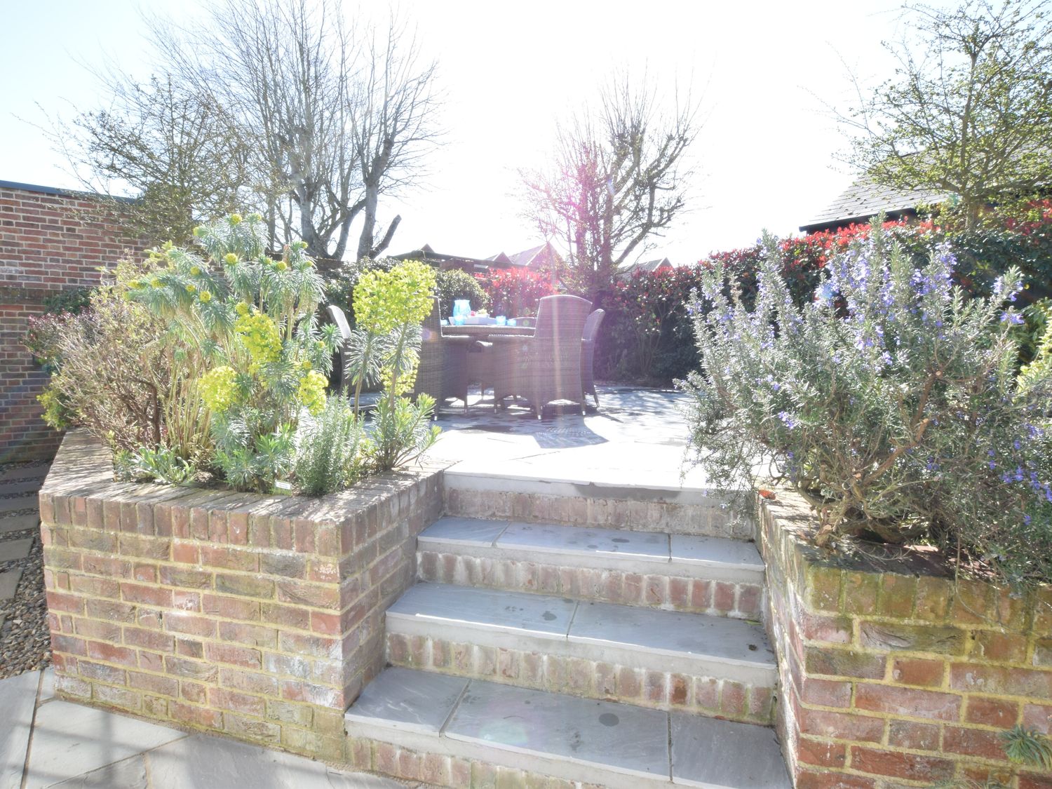 A garden patio with steps leading to a seating area with a table and chairs surrounded by plants and brick walls at Seashells Aldeburgh in Aldeburgh