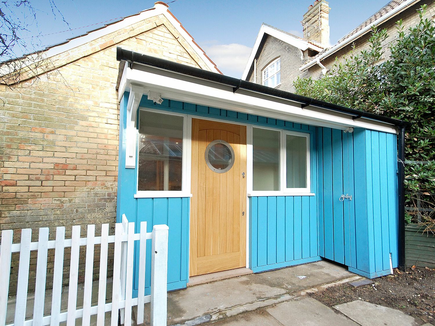 A small blue wooden building with a round window door and white fence at Garden Cottage Southwold