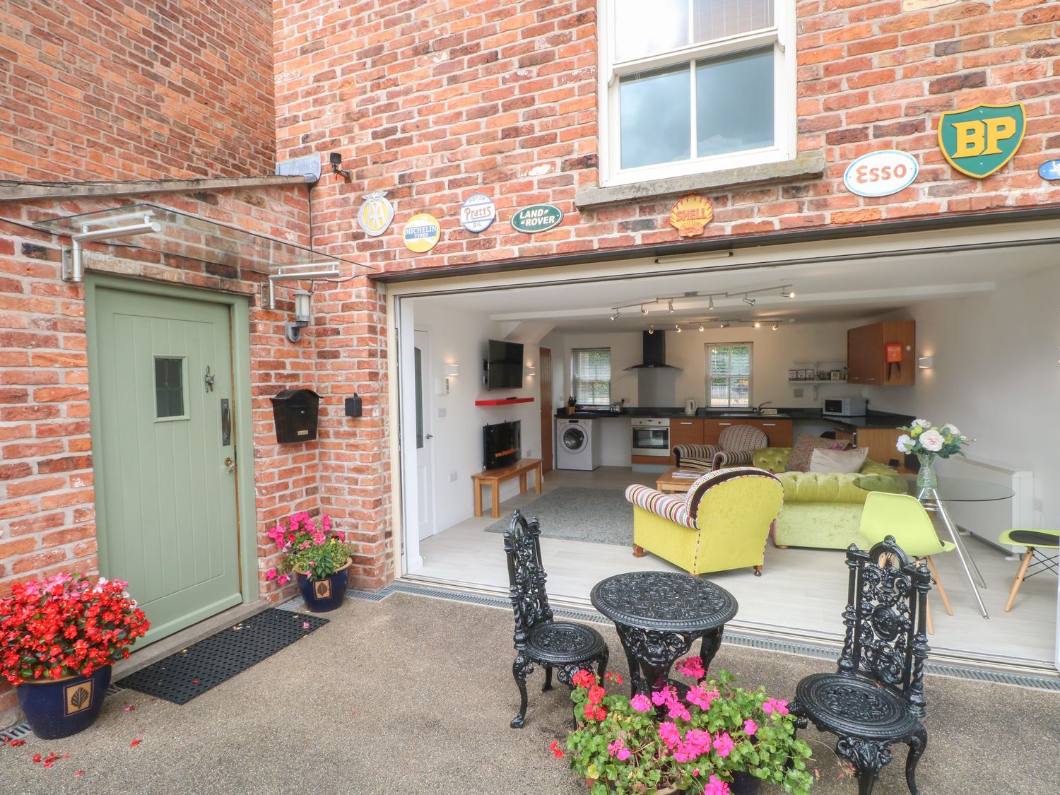 An outdoor patio with black metal table and chairs and pink flowers in pots next to a green door and an open living room with kitchen at The Lodge at Cross Cottage in Holt North Wales