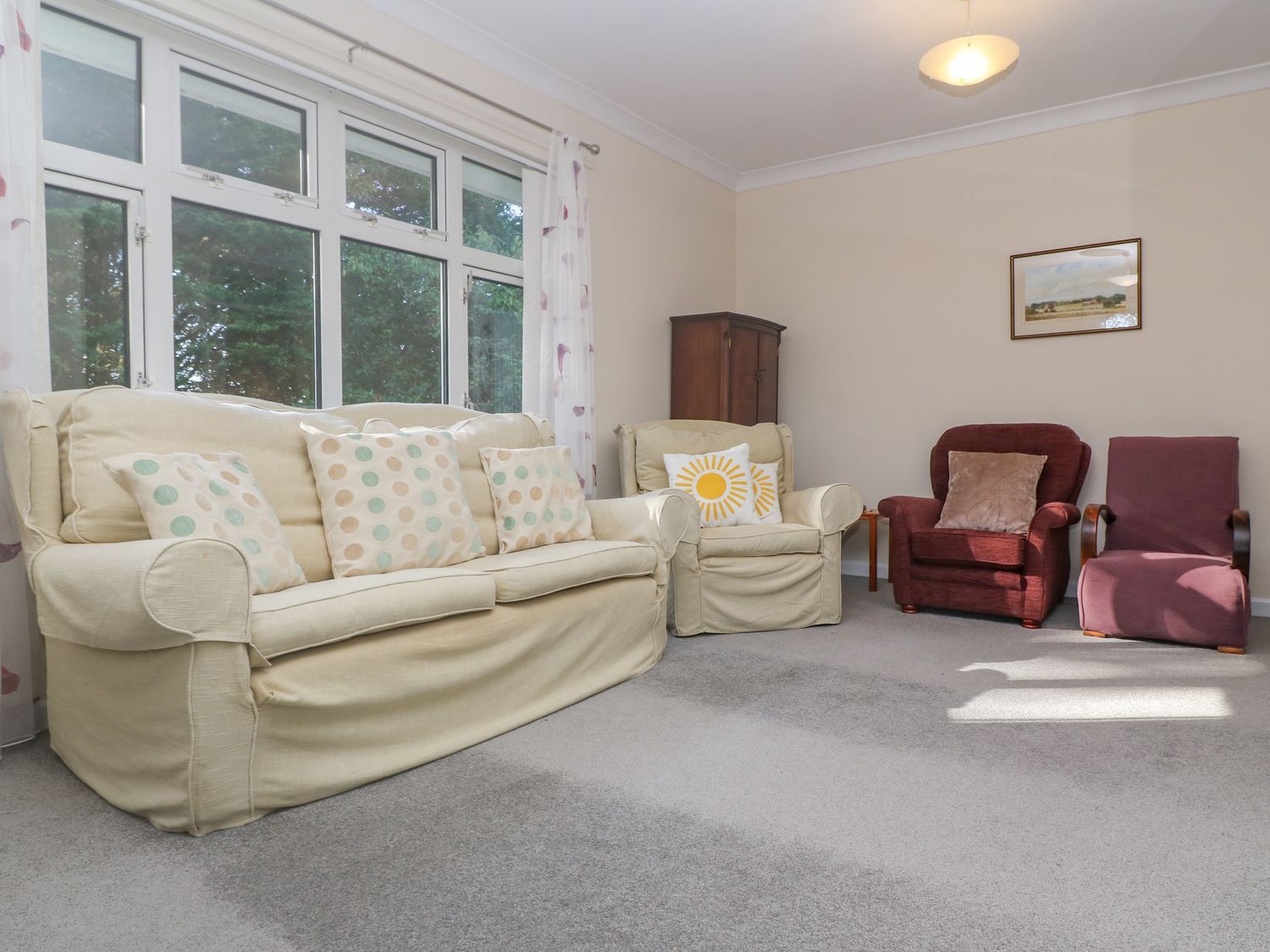 A living room with a beige sofa and armchair with patterned cushions two red armchairs a wooden cabinet and a framed picture on the wall at Yngywidden in Rosudgeon