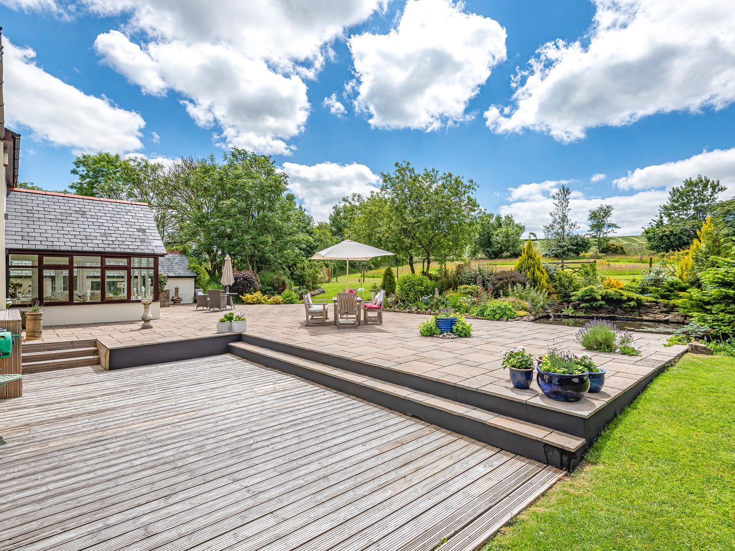 A backyard with a wooden deck and stone patio with outdoor furniture and plants at Waterland Farmhouse in Bradworthy