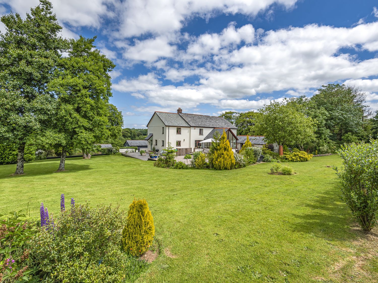 A house with a lawn and trees under a blue sky at Waterland Farmhouse in Bradworthy