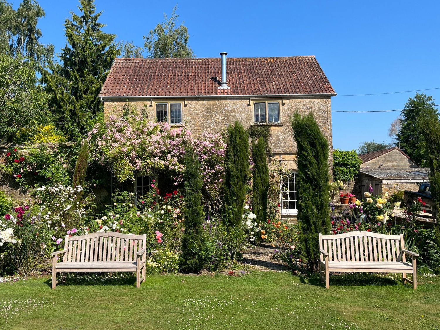 A garden with benches flowers and bushes in front of a stone house with tiled roof at Ham cottage East Lambrook near South Petherton