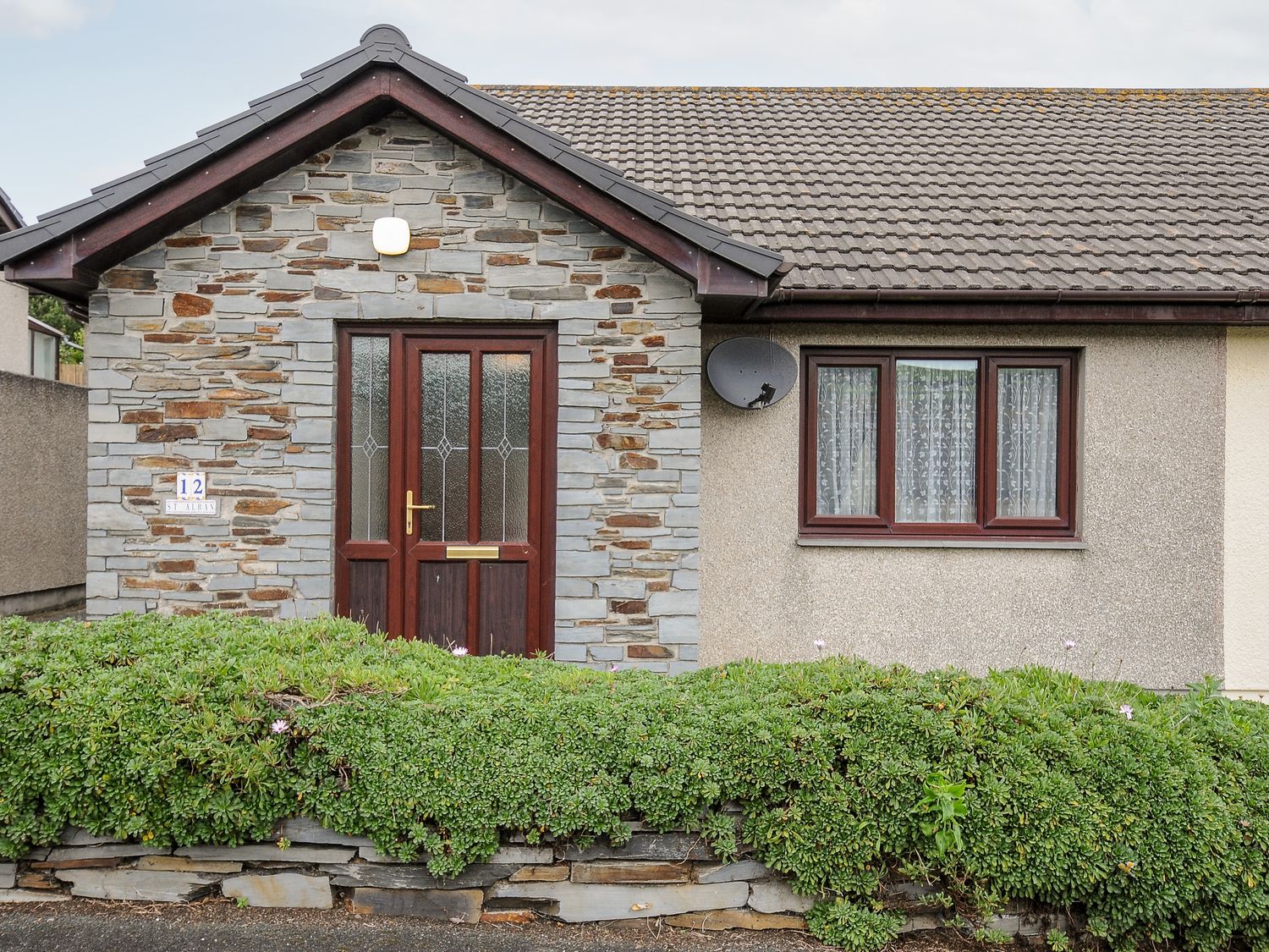 The exterior of a single-story house with a stone entrance door and a window with curtains at St Albans in Boscastle