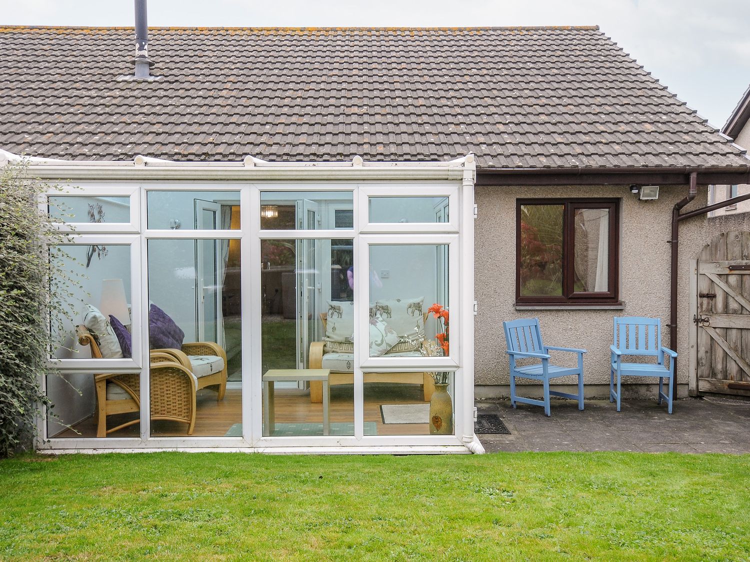 A backyard with a grass lawn two blue wooden chairs and a conservatory with wicker chairs at St Albans in Boscastle