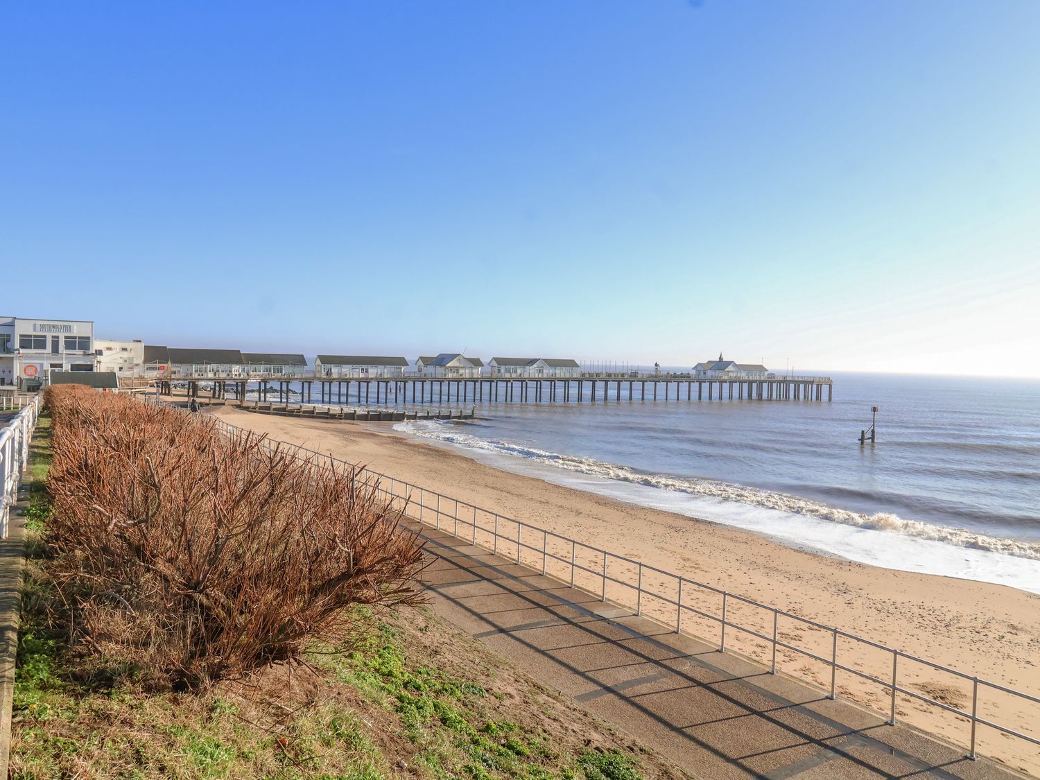 A beach with a pier extending into the sea and a paved walkway with railings alongside it at Seas The Day in Southwold