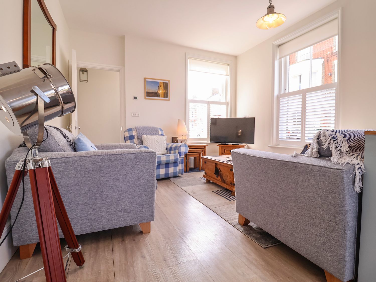 A living room with two grey sofas a blue and white check armchair a wooden coffee table a television and a tripod lamp at Seas The Day in Southwold