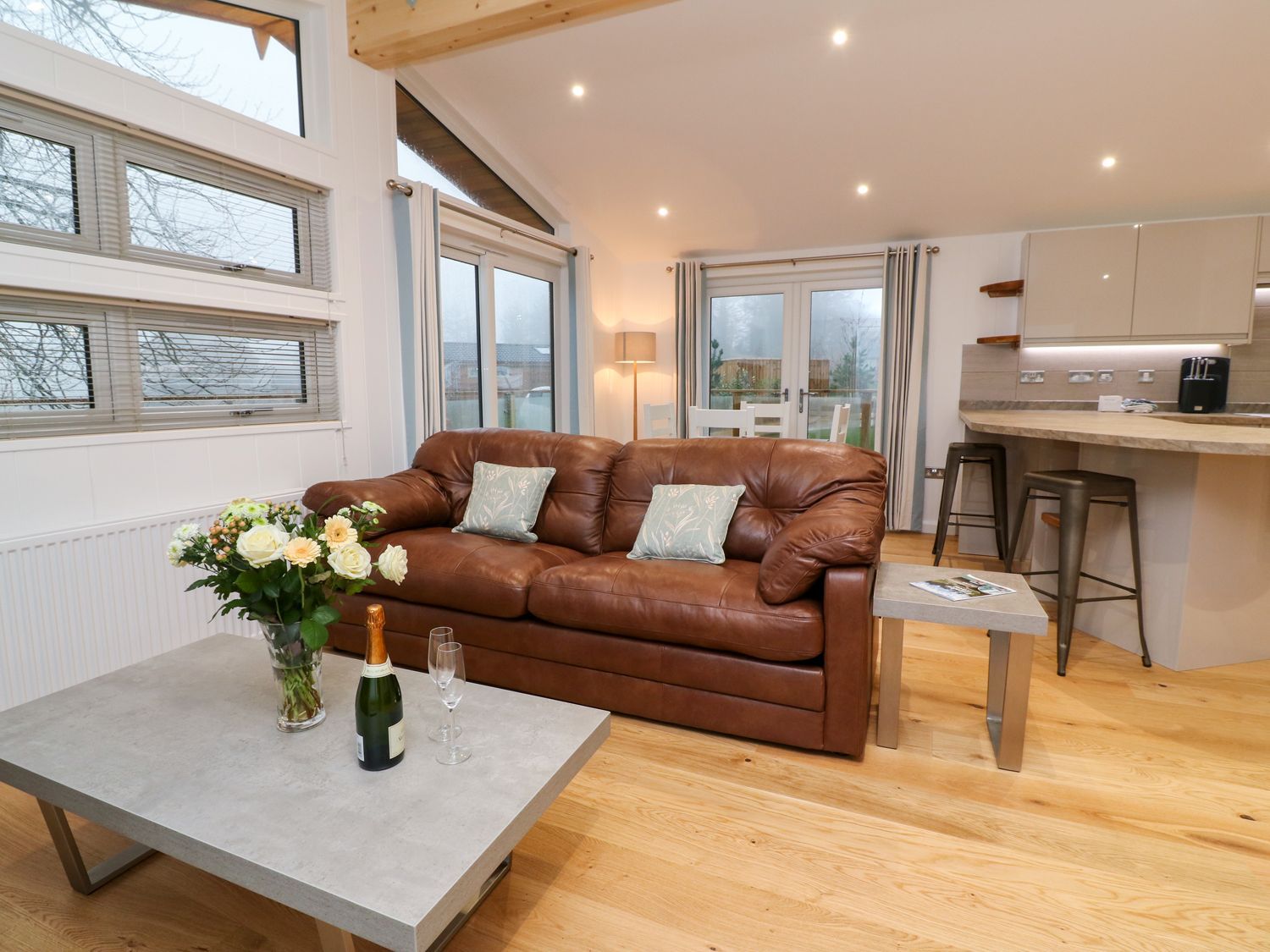 A living room with a brown leather sofa and coffee table with flowers and champagne at 9 Faraway Fields in Dobwalls