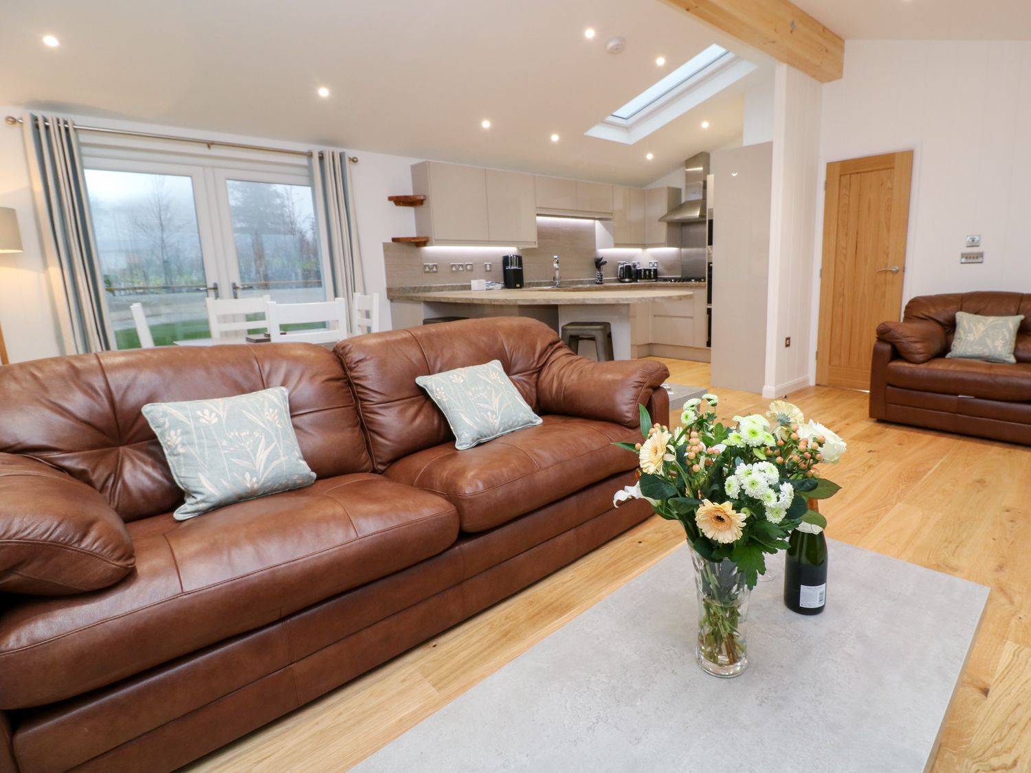 A living room with brown leather sofas a coffee table with flowers and a kitchen in the background at 9 Faraway Fields in Dobwalls