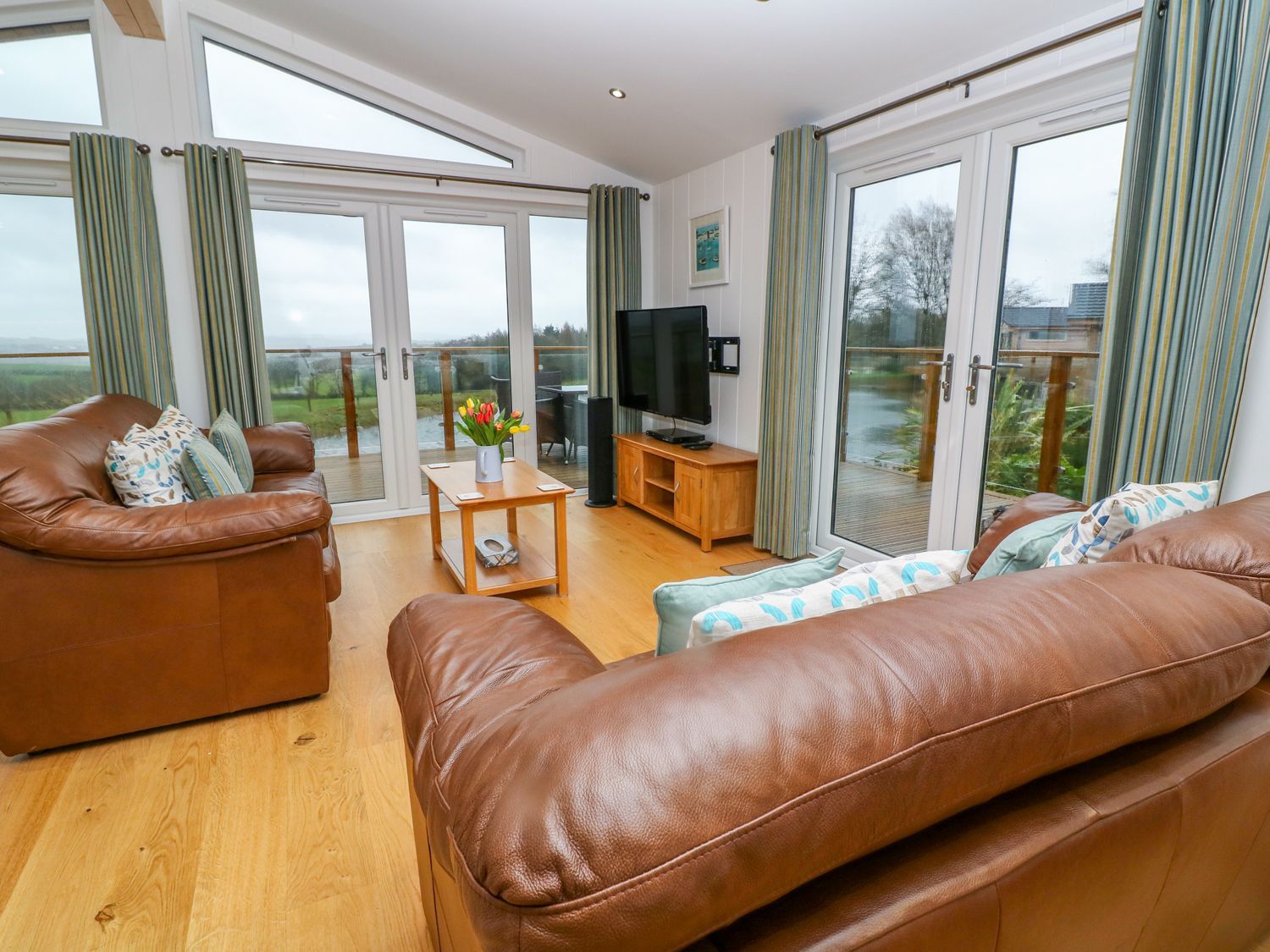 A living room with two brown leather sofas a wooden coffee table a TV on a wooden stand and large windows at 12 Faraway Fields in Dobwalls