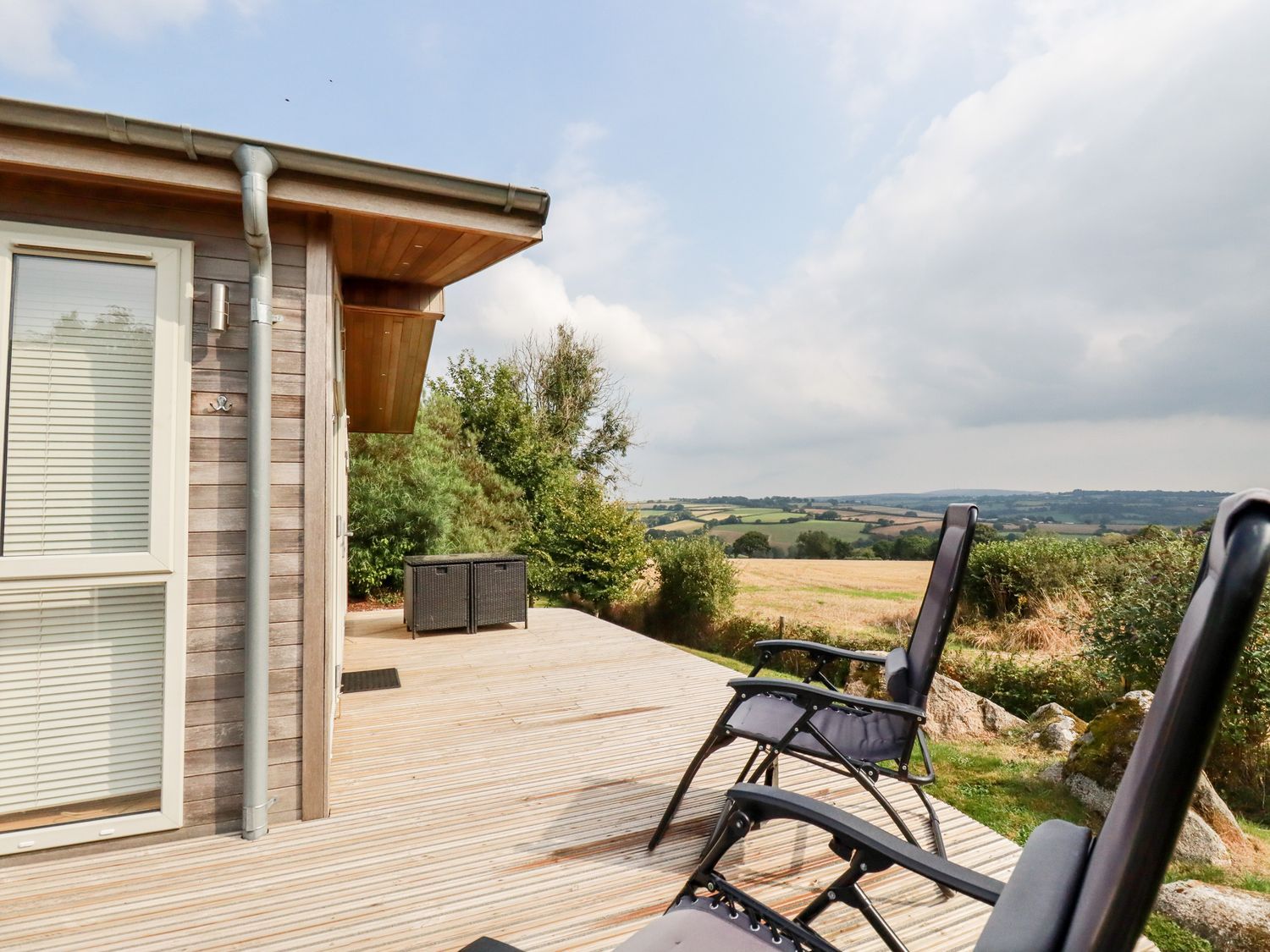 An outdoor wooden deck with two reclining chairs and storage boxes overlooking fields and hills at 11 Horizon View in Dobwalls