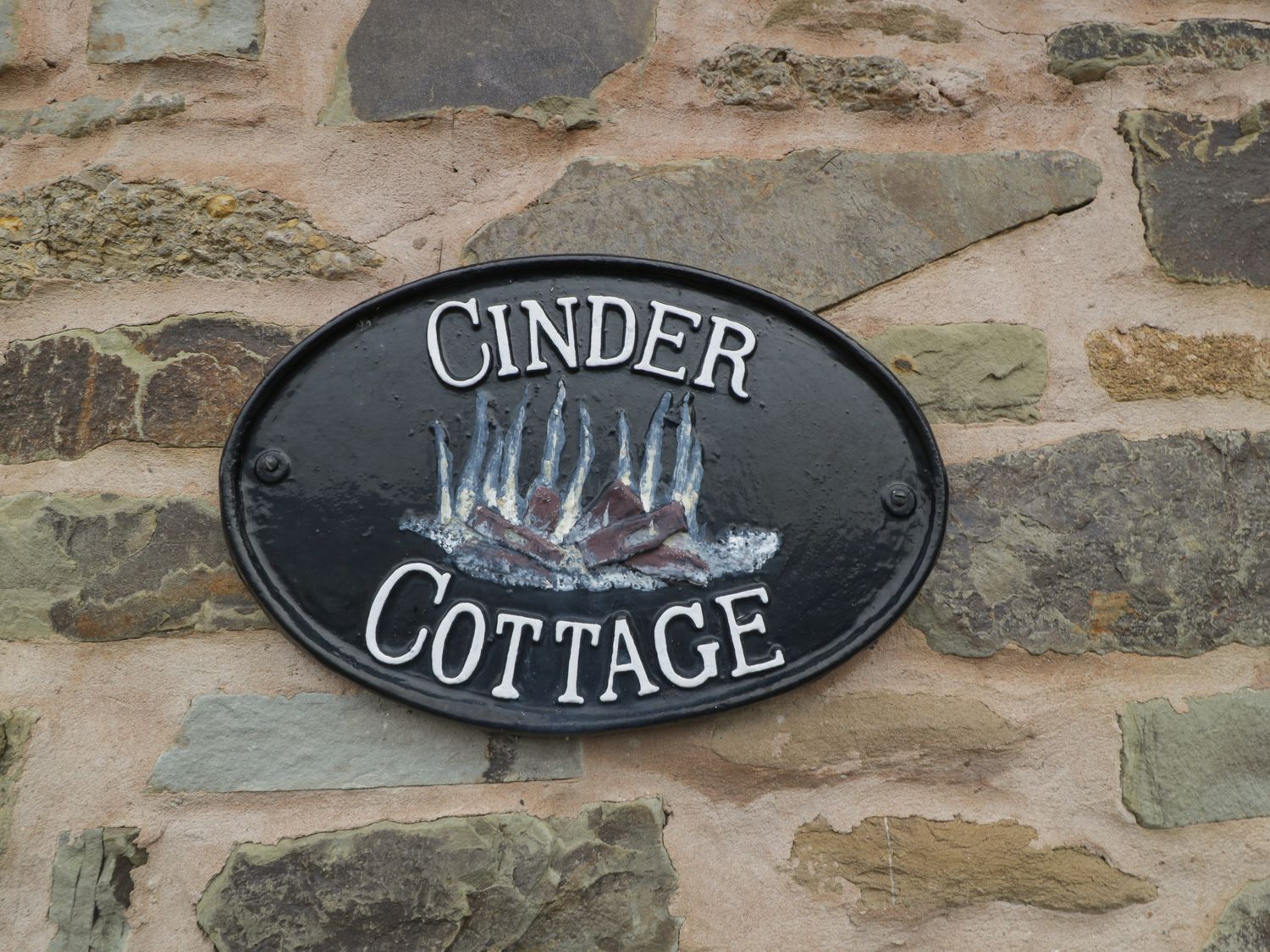 A black oval sign with the words Cinder Cottage and a fire illustration on a stone wall at Cinder Cottage in Stanford Bishop near Bromyard