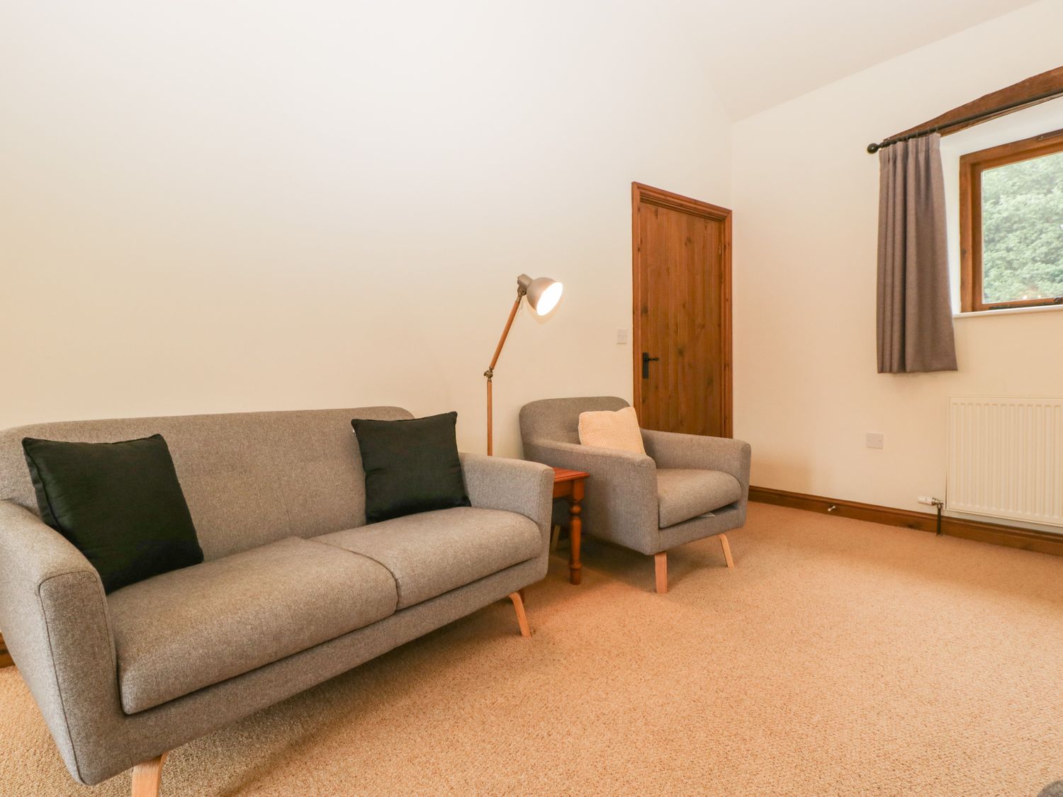 A living room with a grey sofa, matching armchair, wooden side table, floor lamp, and window with curtain at Cinder Cottage Stanford Bishop near Bromyard