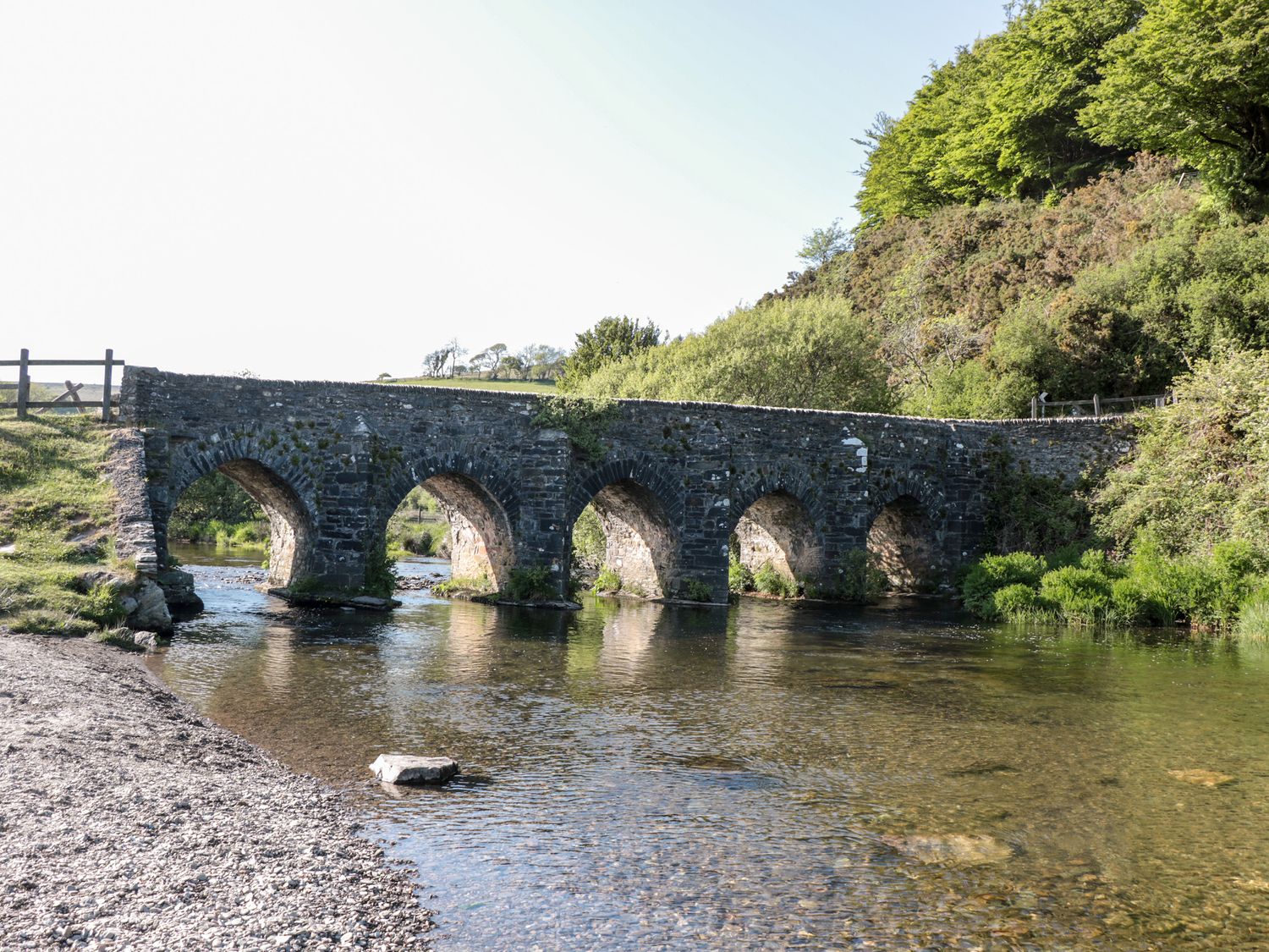 A stone arch bridge over a shallow river with trees and bushes on the riverbank at The Pound in Porlock