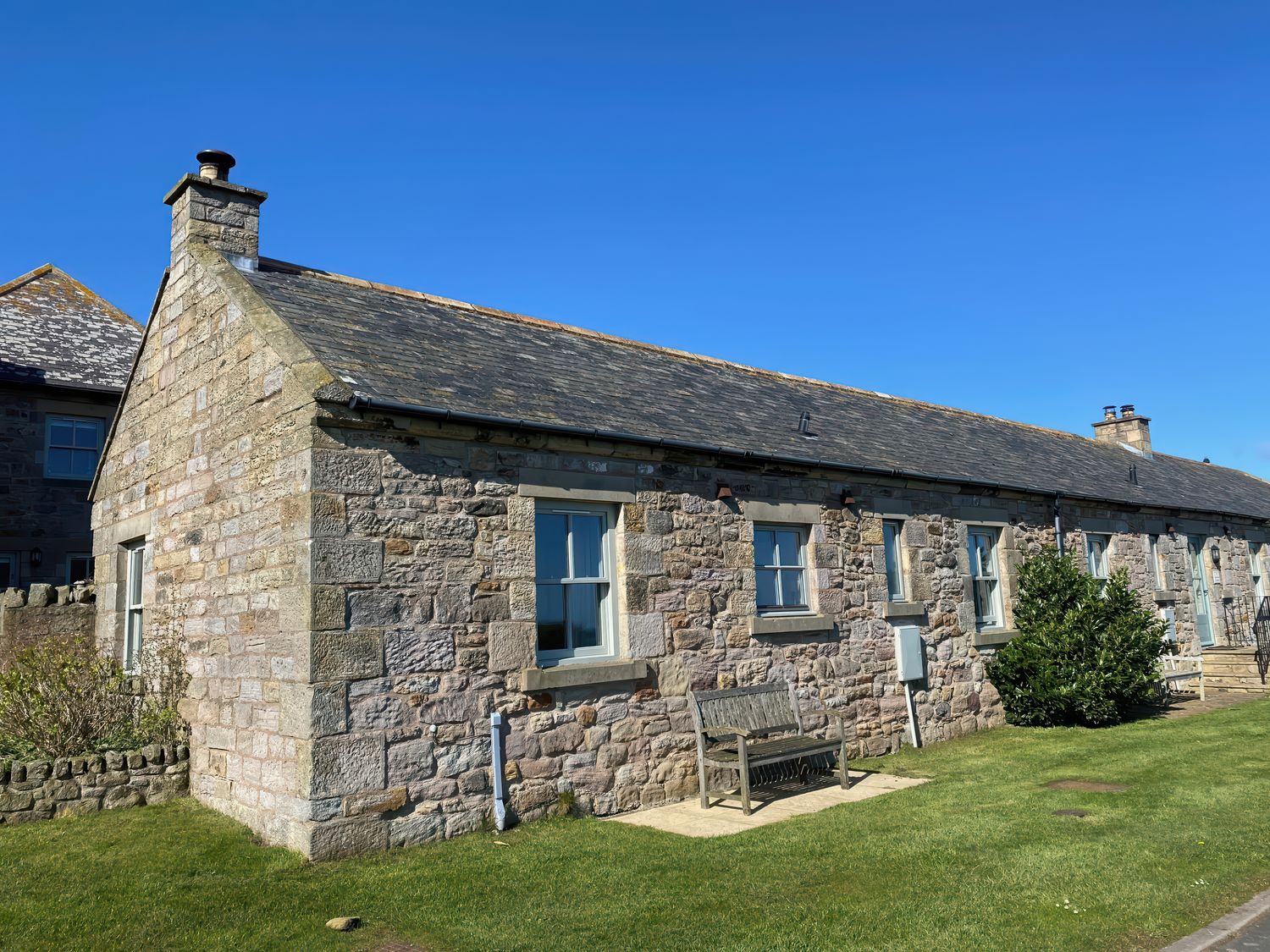 A stone cottage with windows a wooden bench and a small bush on a lawn at Beachward in Embleton