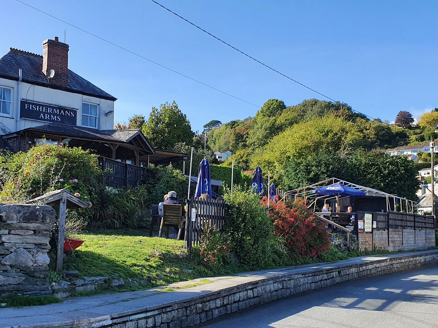 A pub with outdoor seating and umbrellas next to a road surrounded by greenery and trees