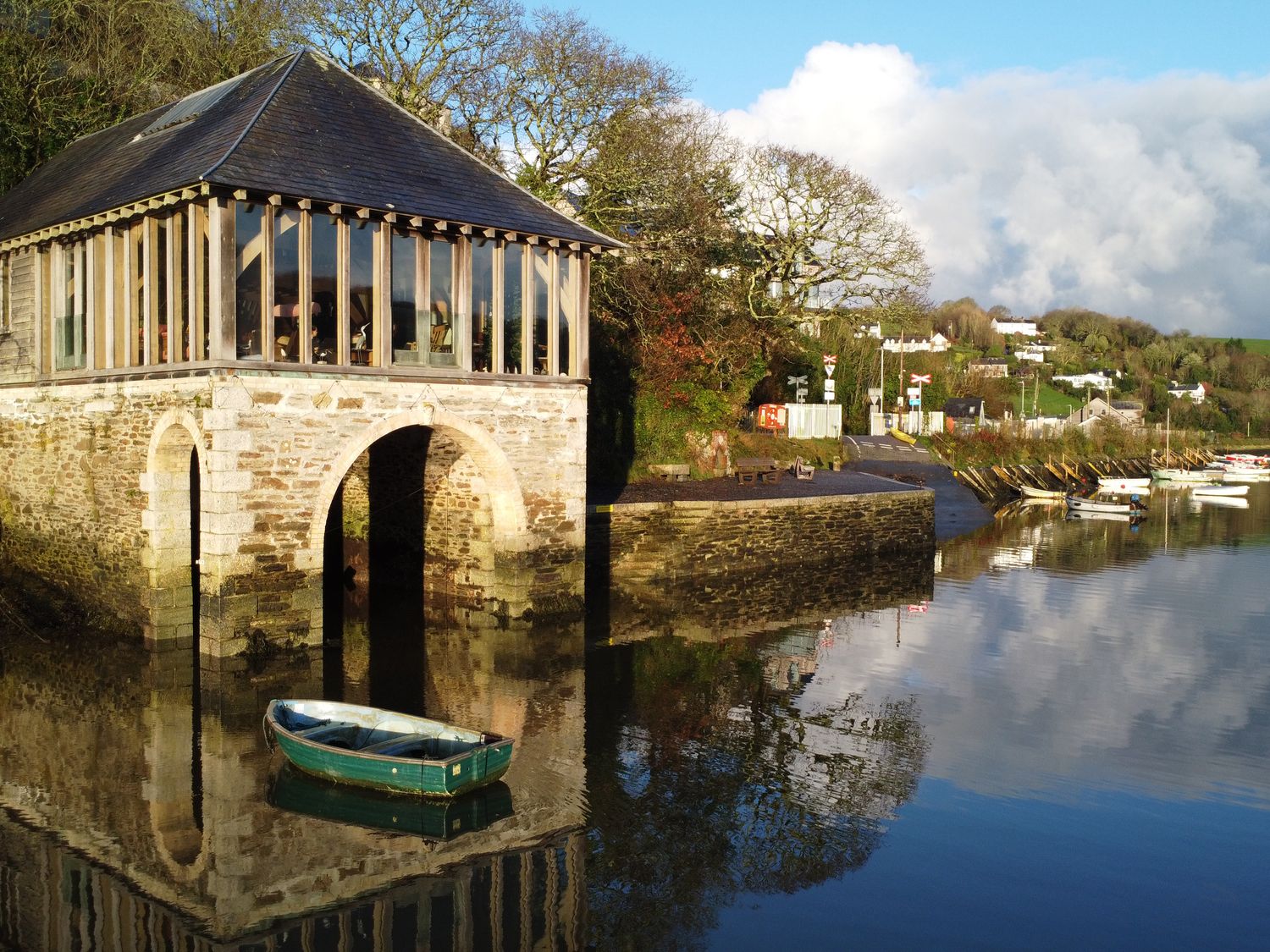 A stone building with arched openings by the water with a small rowboat floating nearby and several boats moored in the background at Oakenhurst in Golant