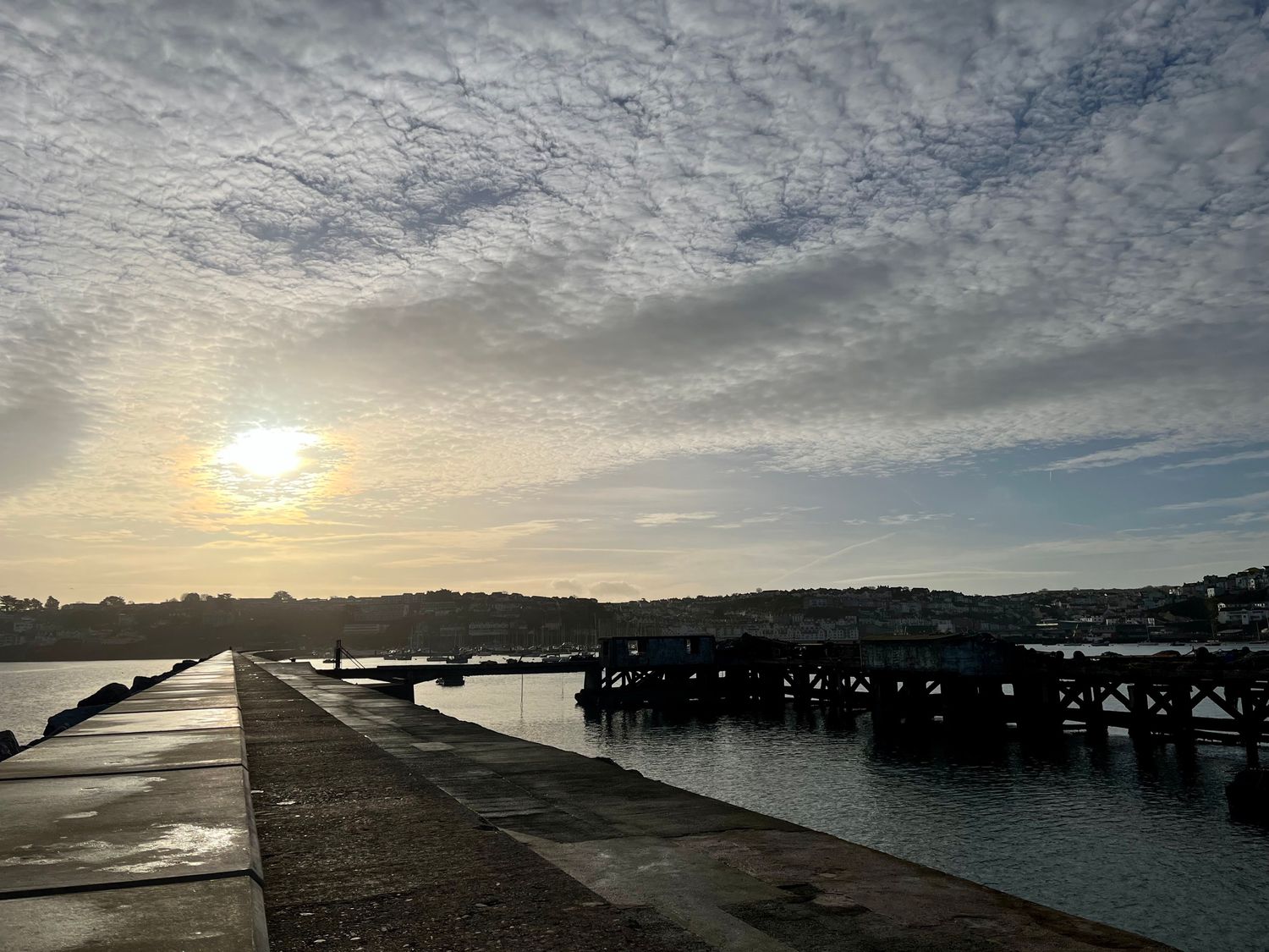 A concrete pier extending into water with a wooden structure and houses on hills in the background at Ingledene in Brixham