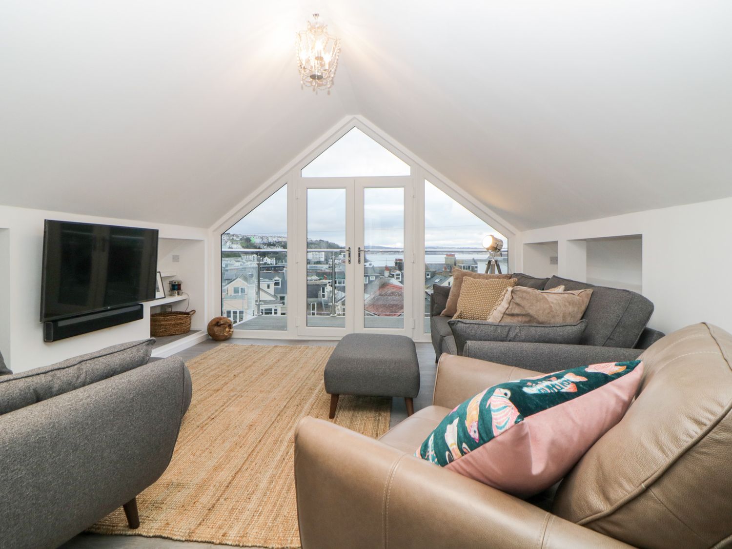 A living room with sofas, an ottoman, a wall-mounted TV, and large windows overlooking rooftops at Ingledene in Brixham