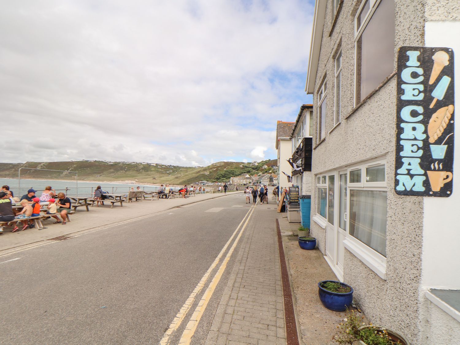 A seaside street with people sitting at picnic tables near the water and an ice cream shop sign at Harbour Studio in Sennen Cove
