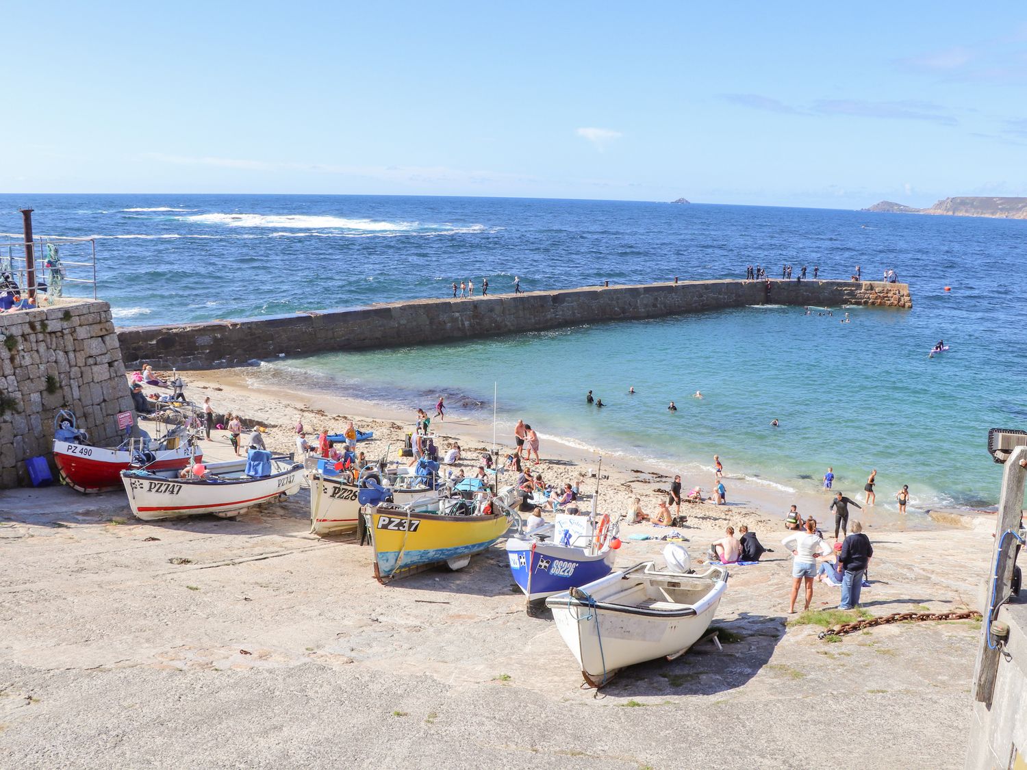 A small beach area with several boats on a ramp and people swimming and walking near a sea wall at Harbour Studio in Sennen Cove