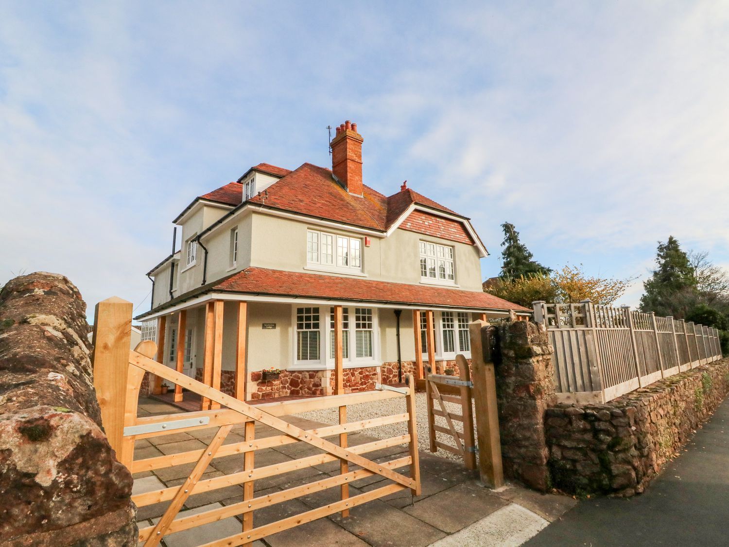 A detached two-story house with a red tiled roof behind a wooden gate and stone wall at Westholme Lodge in Minehead