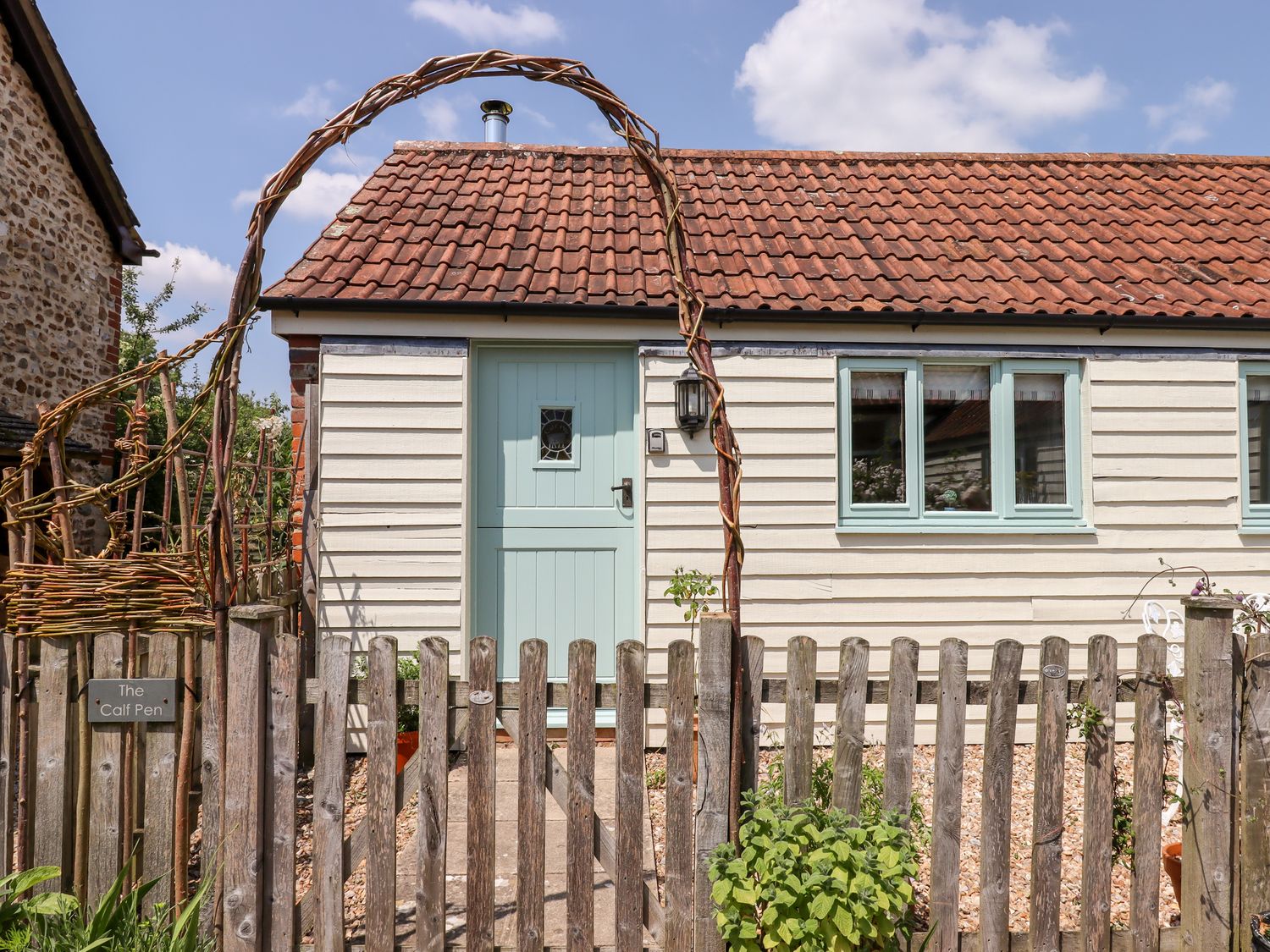 A small house with a light blue door and windows behind a wooden fence with a woven arch at The Calf Pen in Colyton