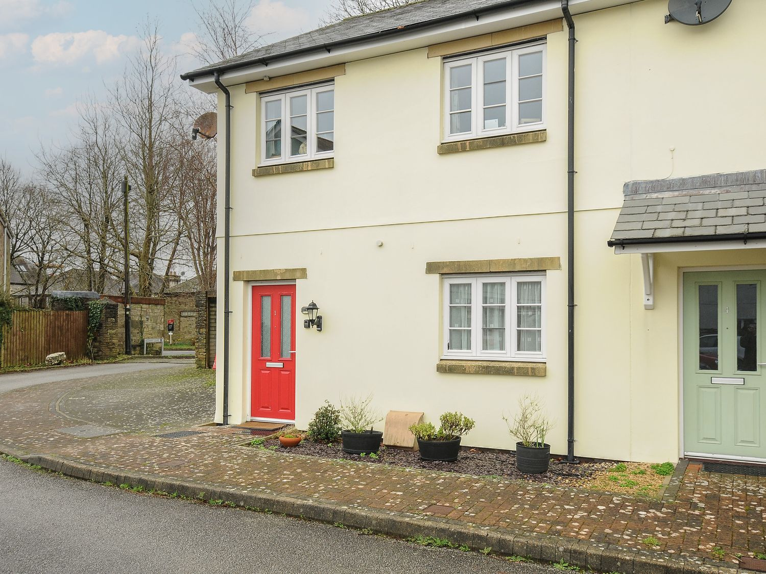 The exterior of a two-story house with a red front door several potted plants on a paved area and a road in front at 1 Bethany Court Bodmin