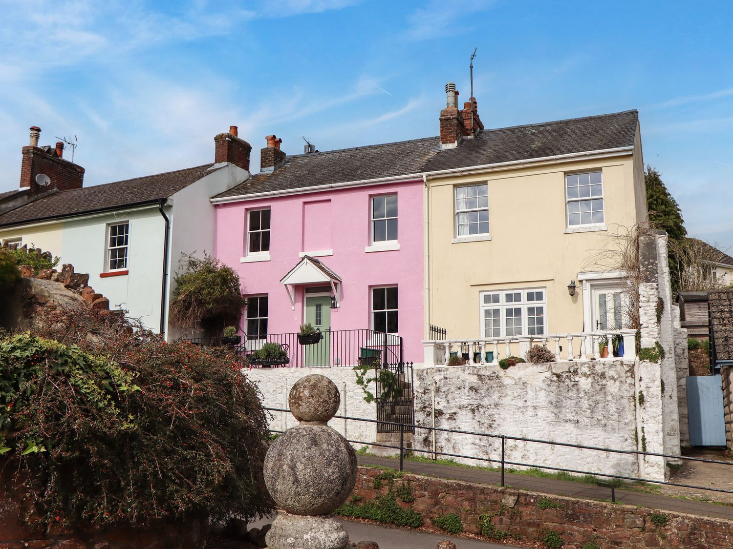 A street view of three attached houses with pastel colored walls and a stone wall in front at Rose Cottage in Bishopsteignton