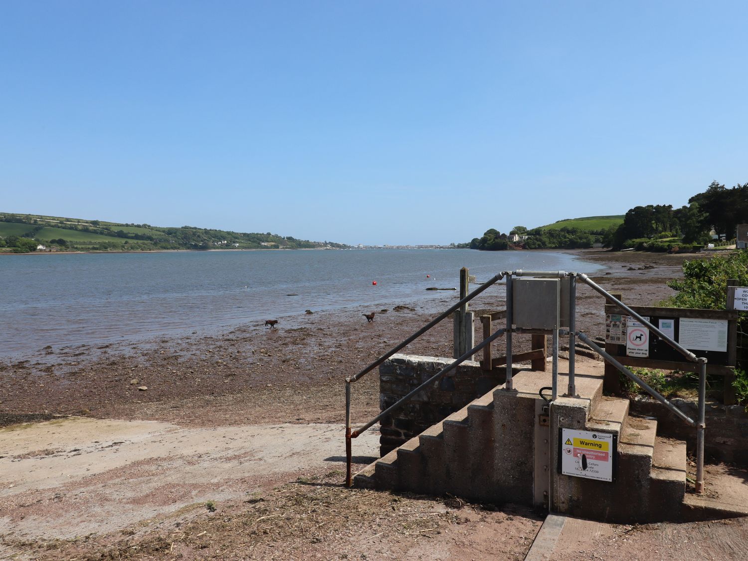 A riverside area with a concrete staircase and metal railing leading to a muddy shore with grassy hills in the background at Rose Cottage in Bishopsteignton